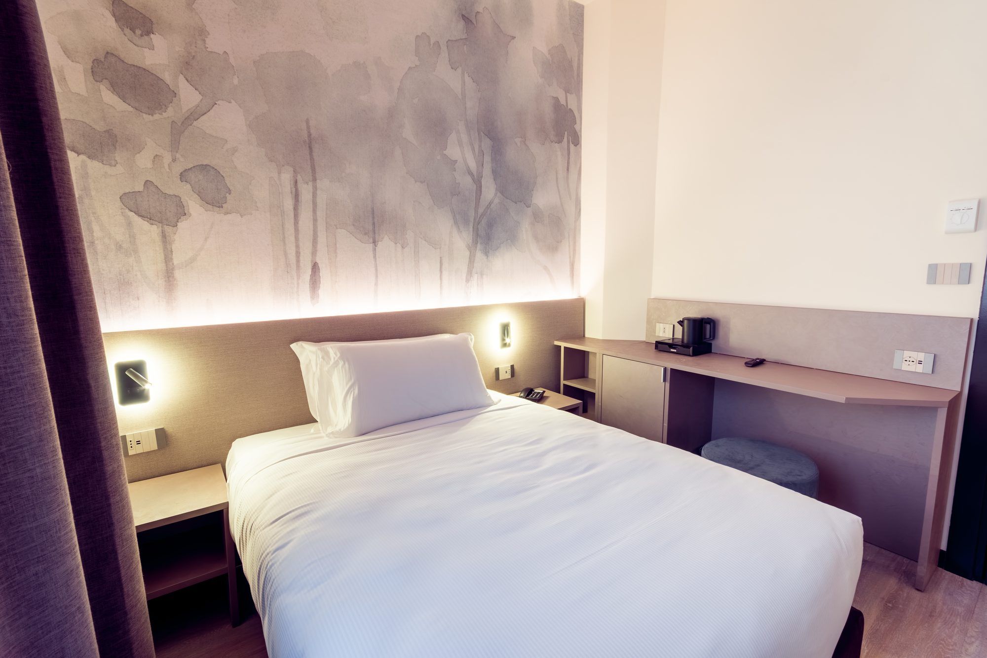 A hotel room with a bed, bedside tables, a desk, and a wall-mounted light fixture.