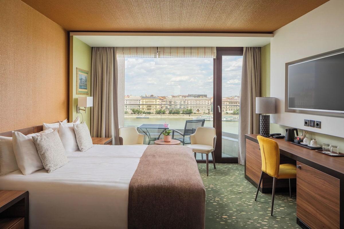 A hotel room with a bed, a desk, a chair, a TV, and a balcony with a view of buildings.