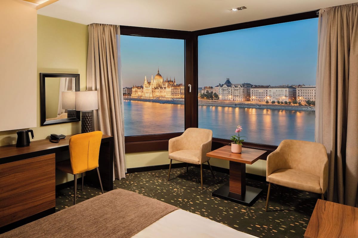 A hotel room with a large window offering a view of a historic building and a river. There is a desk with a lamp, a chair, and a small table with a flower vase.