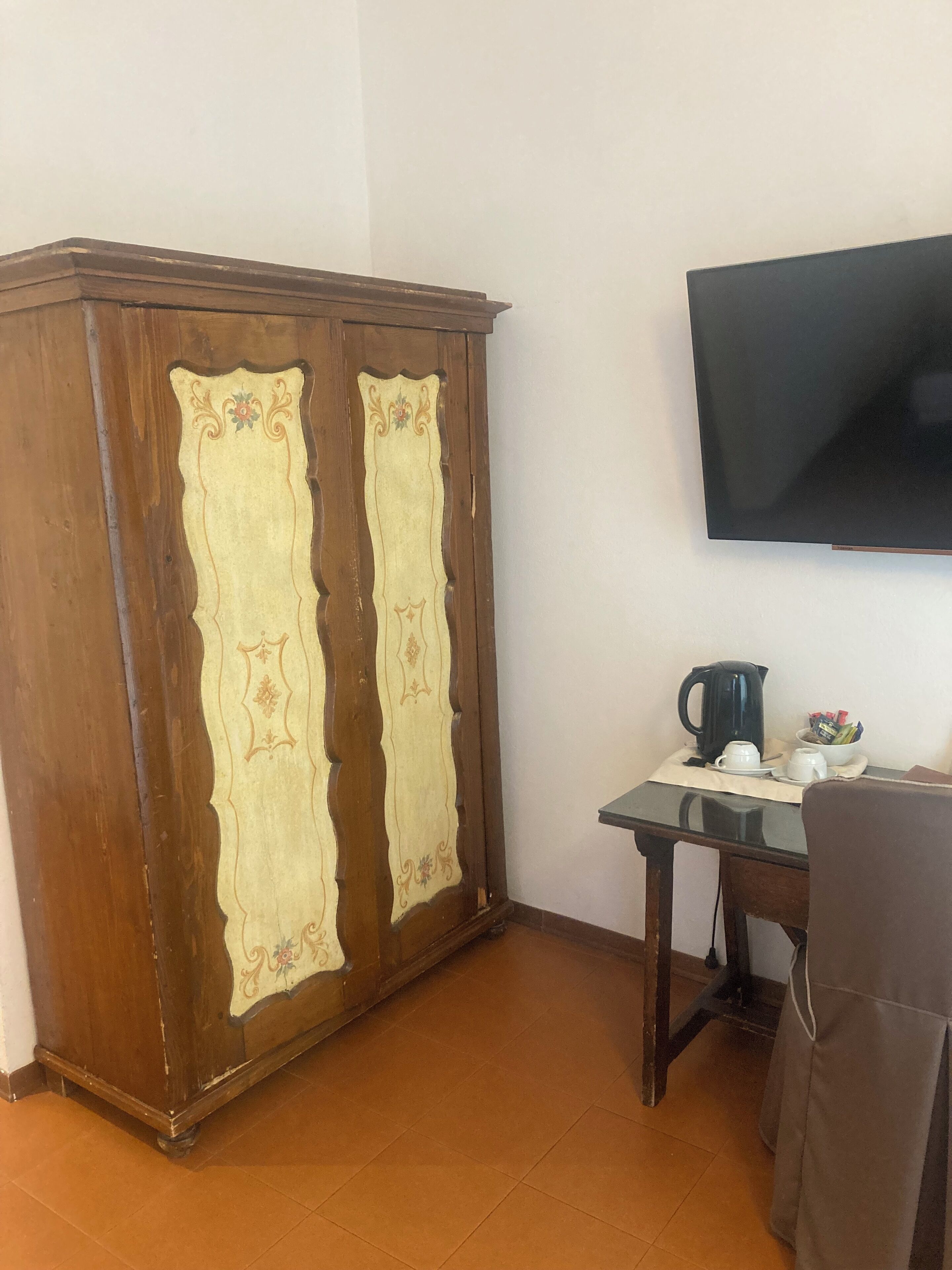 A room with a wooden wardrobe, a small table with a kettle and cups, and a flat-screen TV mounted on the wall.