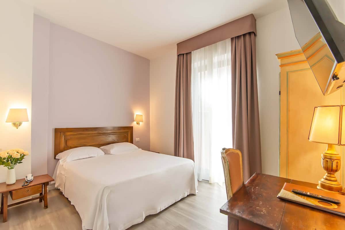 Habitación de hotel con cama, lámparas de noche, escritorio con silla y ventana con cortinas.