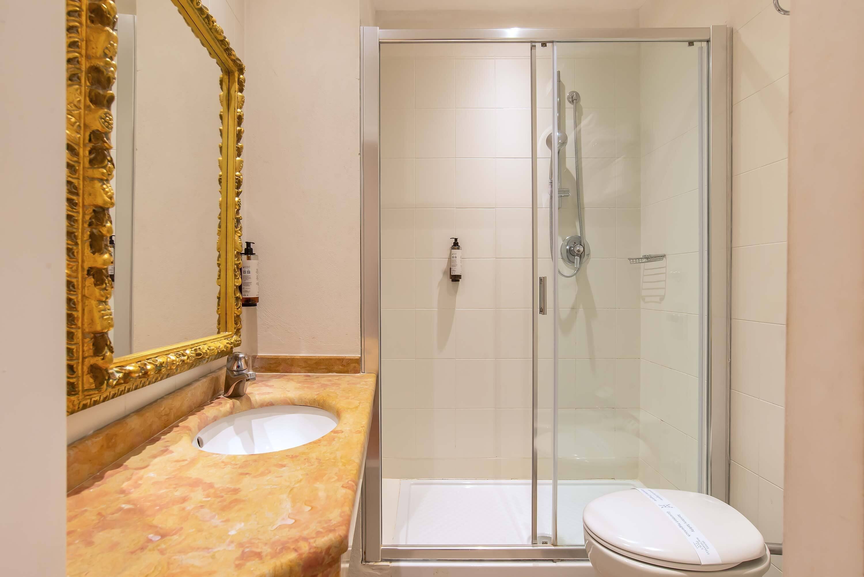 A bathroom with a marble sink, a gold-framed mirror, a glass-enclosed shower, and a white toilet.