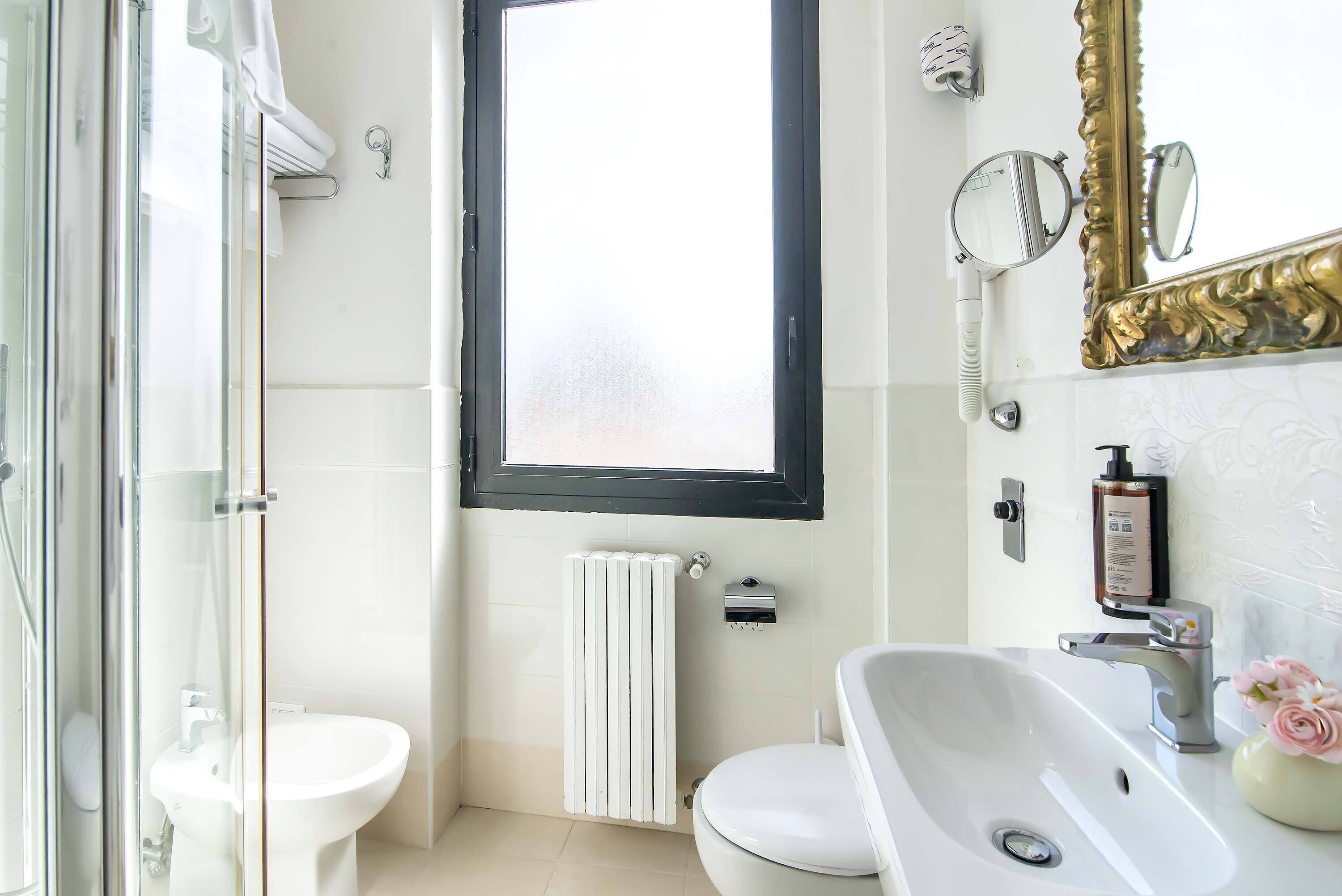 Une salle de bain avec une douche à parois de verre, des toilettes, un bidet, un lavabo avec un miroir et une fenêtre située au-dessus des toilettes.
