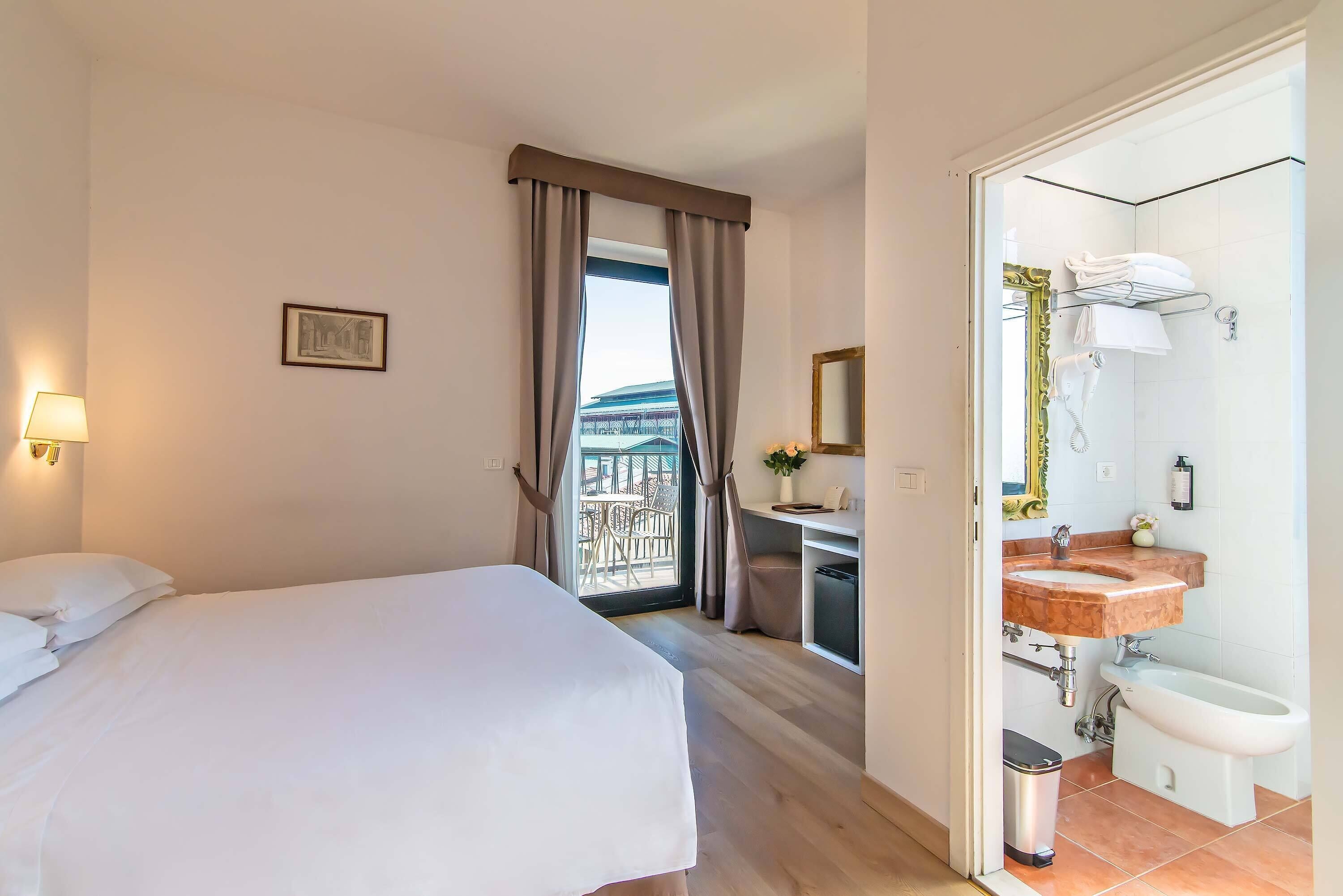 A hotel room with a bed, a desk with a chair, a bathroom with a sink and bidet, and a view of a balcony.
