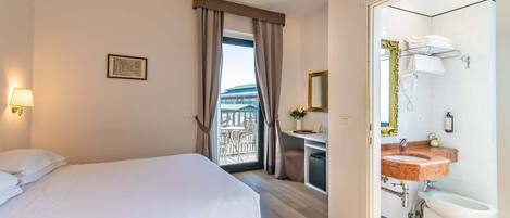 A hotel room with a bed, a desk with a chair, a bathroom with a sink and bidet, and a view of a balcony.