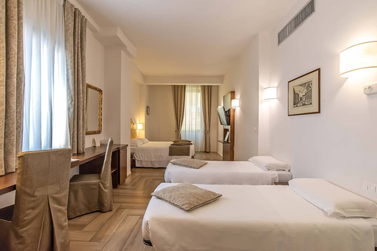 A hotel room with two beds, a desk, and a chair. There are curtains, a mirror, and wall-mounted lights.