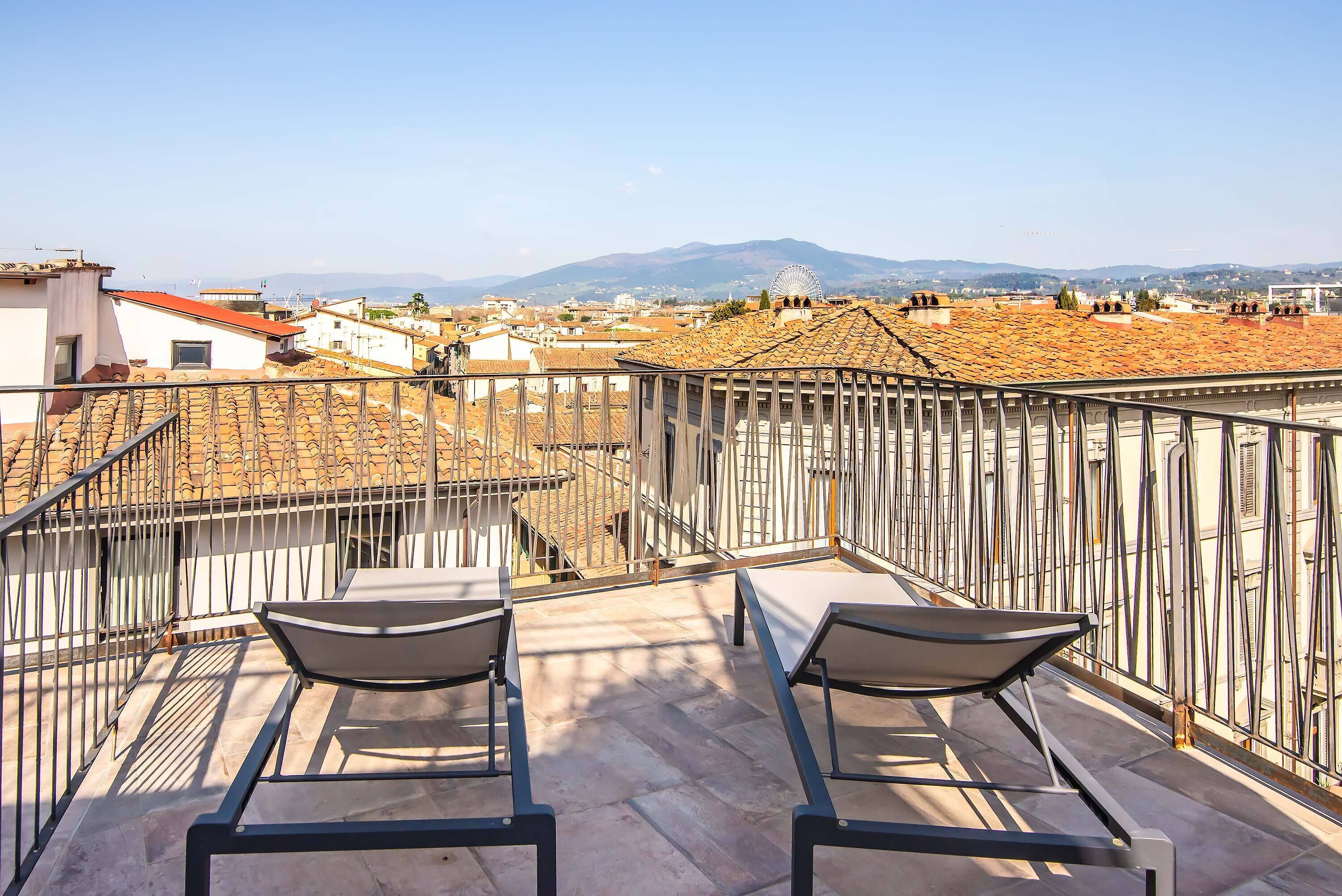 A rooftop terrace with two chairs, a metal railing, and a view of a cityscape with buildings and a mountain in the distance.