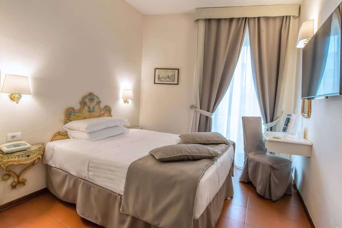 A hotel room with a bed, bedside lamps, a desk with a telephone, a chair, a small table, a mirror, and a window with curtains.