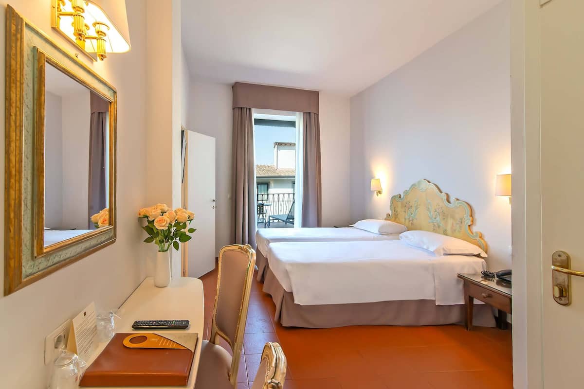 A hotel room with a bed, bedside table, lamp, mirror, desk with a vase of flowers, and a view of the outside.