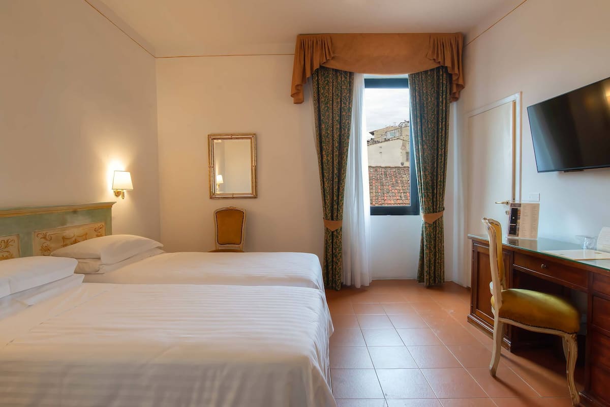 A hotel room with two beds, a desk with a chair, a TV, and a window with curtains.