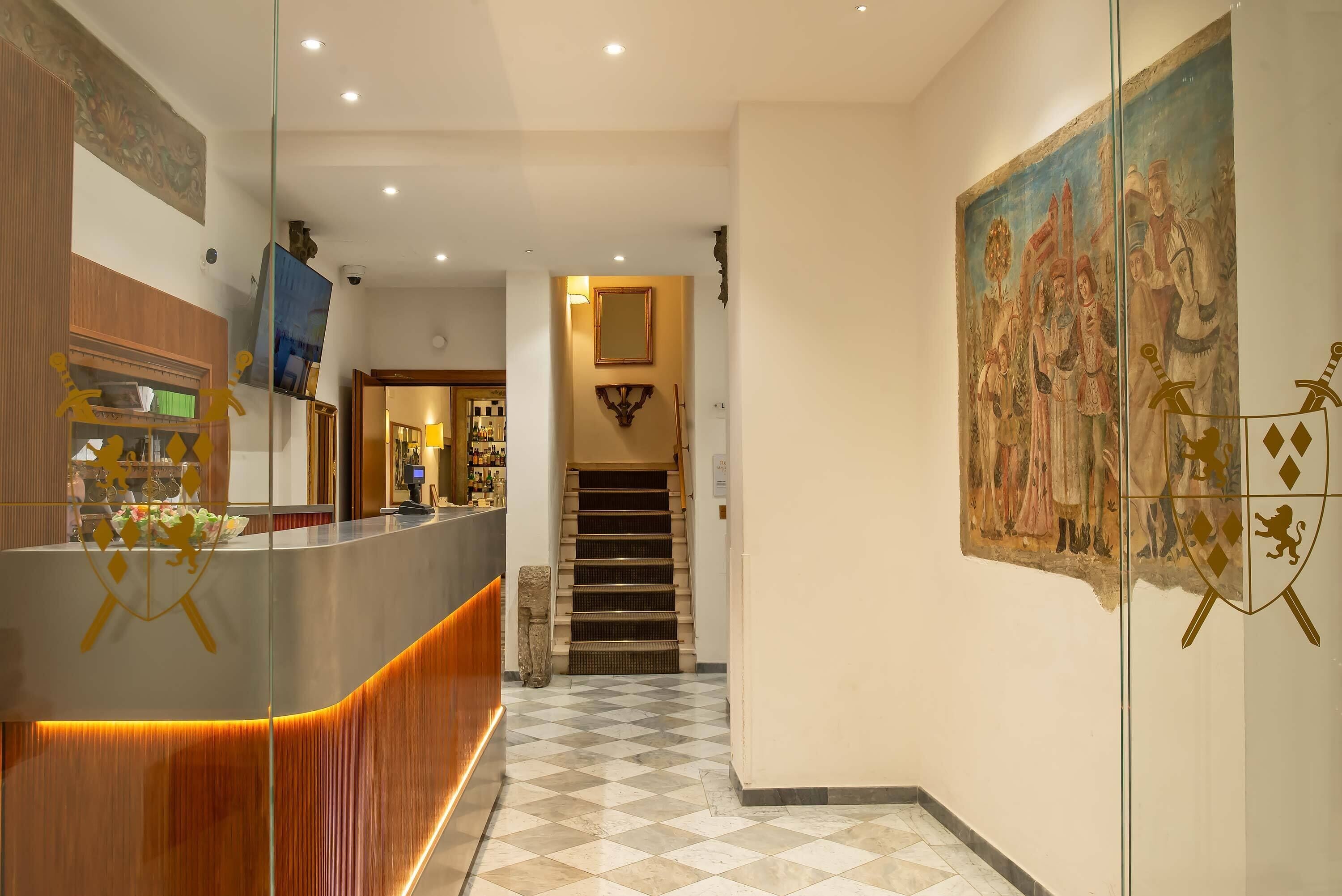 A hotel lobby with a reception desk, a staircase, and a large historical painting on the wall.