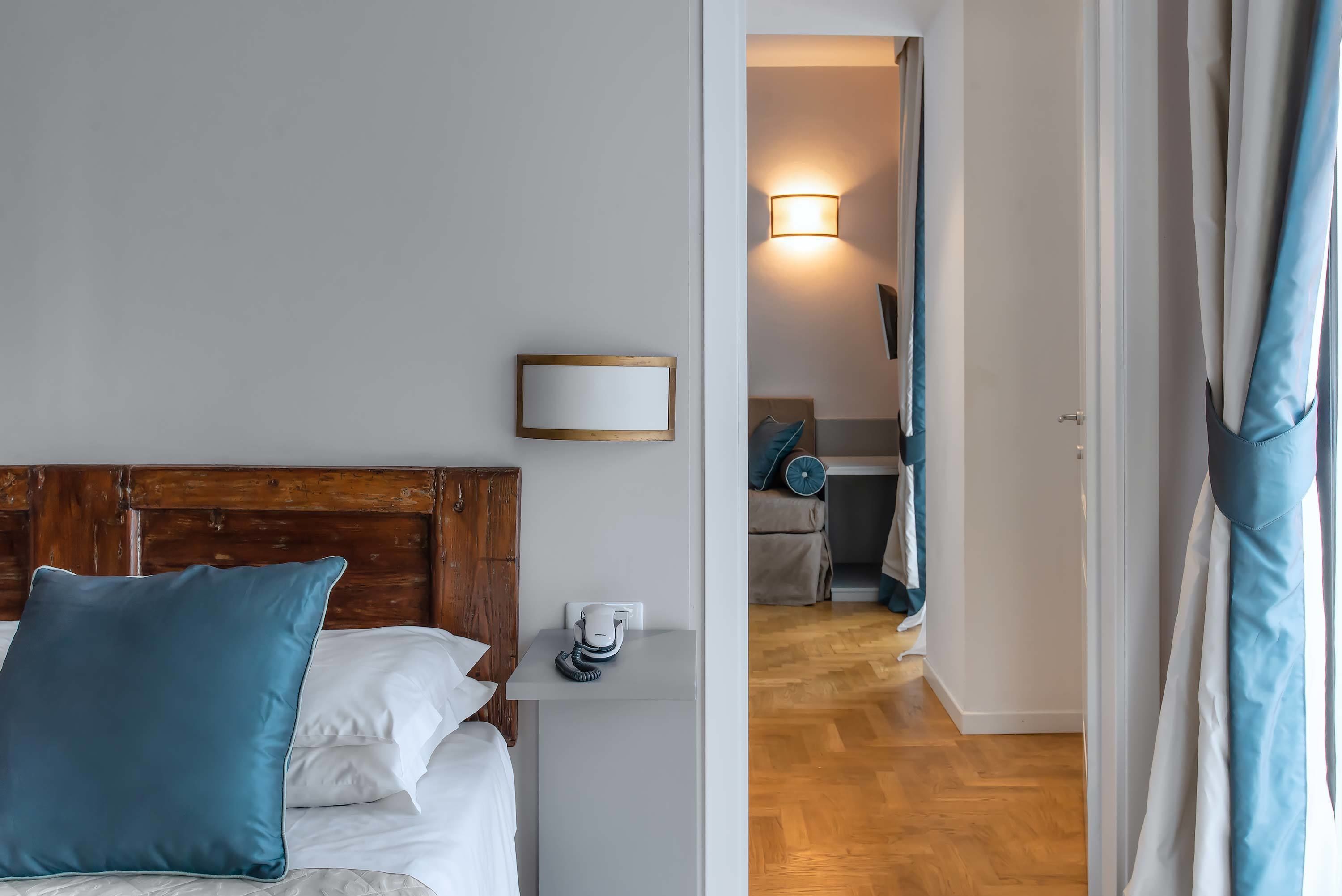 Habitación de hotel con cabecera de madera, mesita de noche con teléfono y vista hacia la habitación contigua.