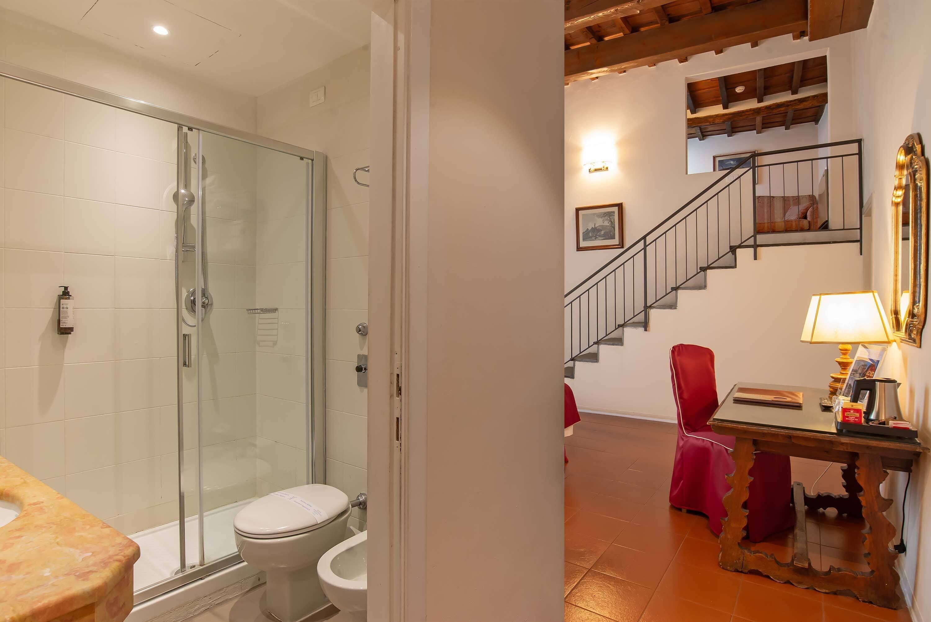 A bathroom with a glass shower enclosure, a toilet, and a sink. There is a staircase leading to an upper level with a red chair and a table with a lamp.
