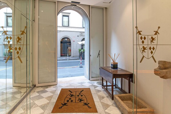 Entrada de hotel con puerta de vidrio, felpudo con escudo, consola de madera y escultura de piedra.
