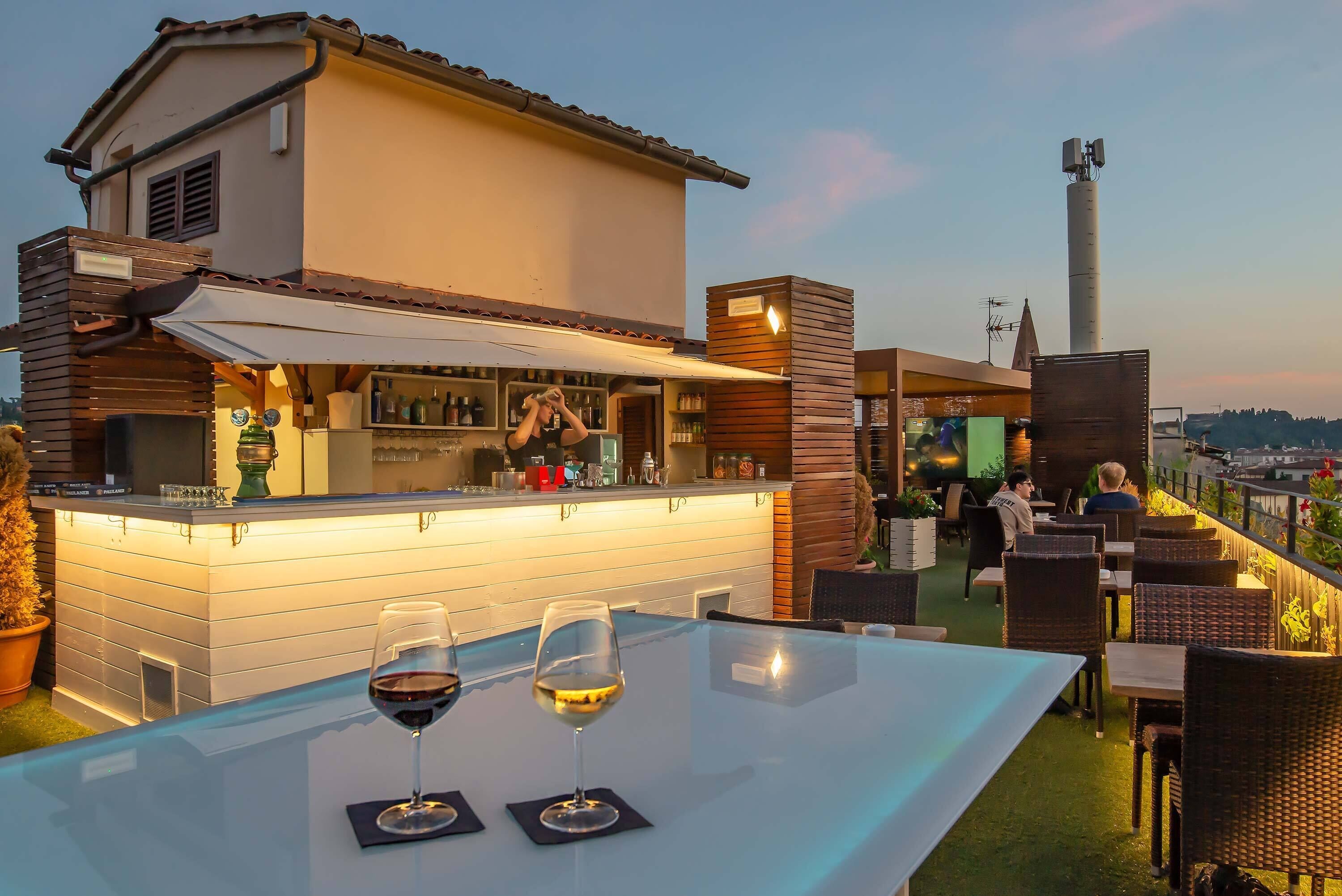 An outdoor bar area with a glass table, two wine glasses, and a bar counter with bottles and glasses.