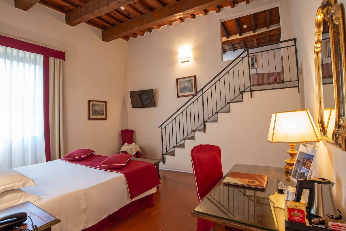 A hotel room with a bed, a desk with a lamp, a chair, a telephone, and a staircase.