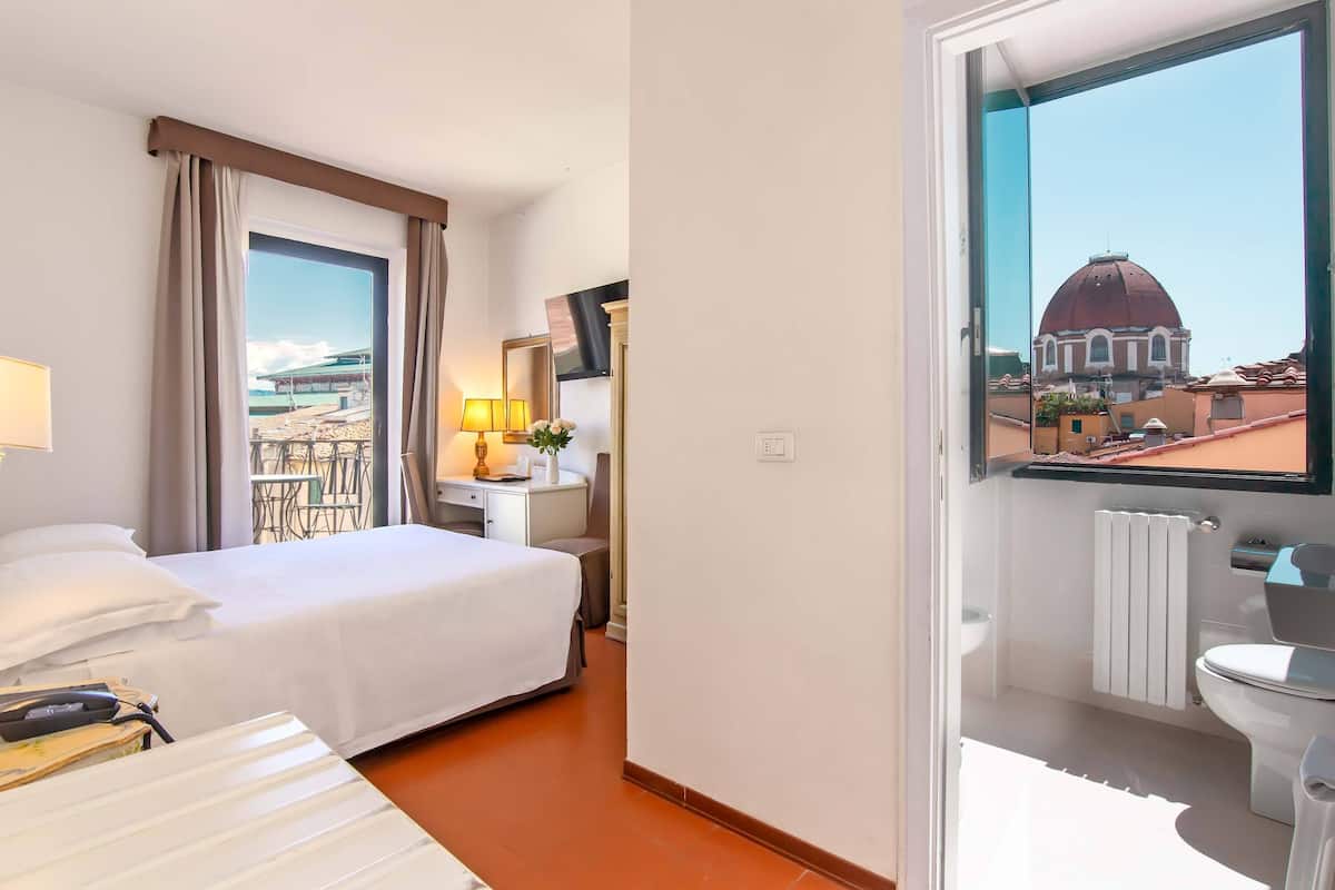 Habitación de hotel con cama, mesita de noche, lámpara y vista a un edificio con cúpula a través de una ventana.