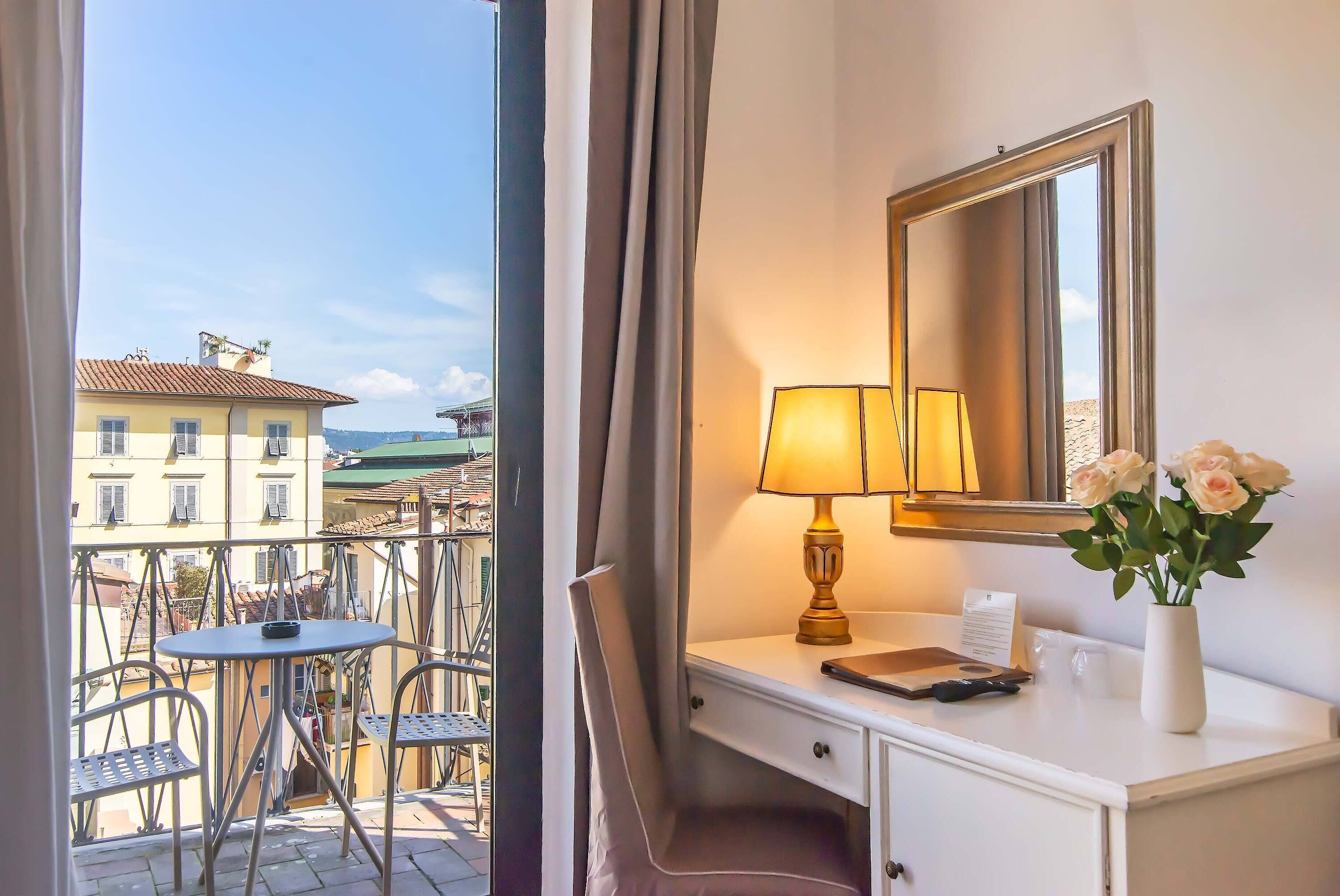 A hotel room with a balcony, a desk with a lamp, a mirror, and a vase with flowers.