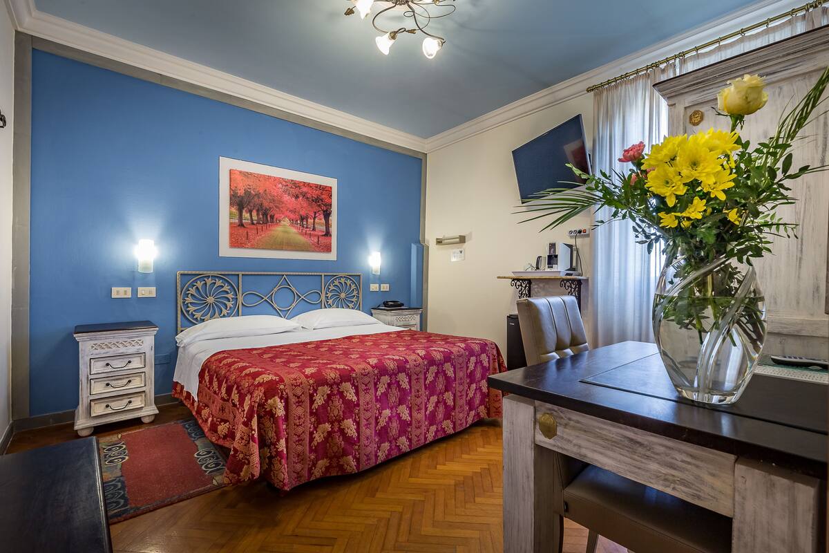 A bedroom with a bed, a desk, a chair, a nightstand, a dresser, and a vase with flowers.