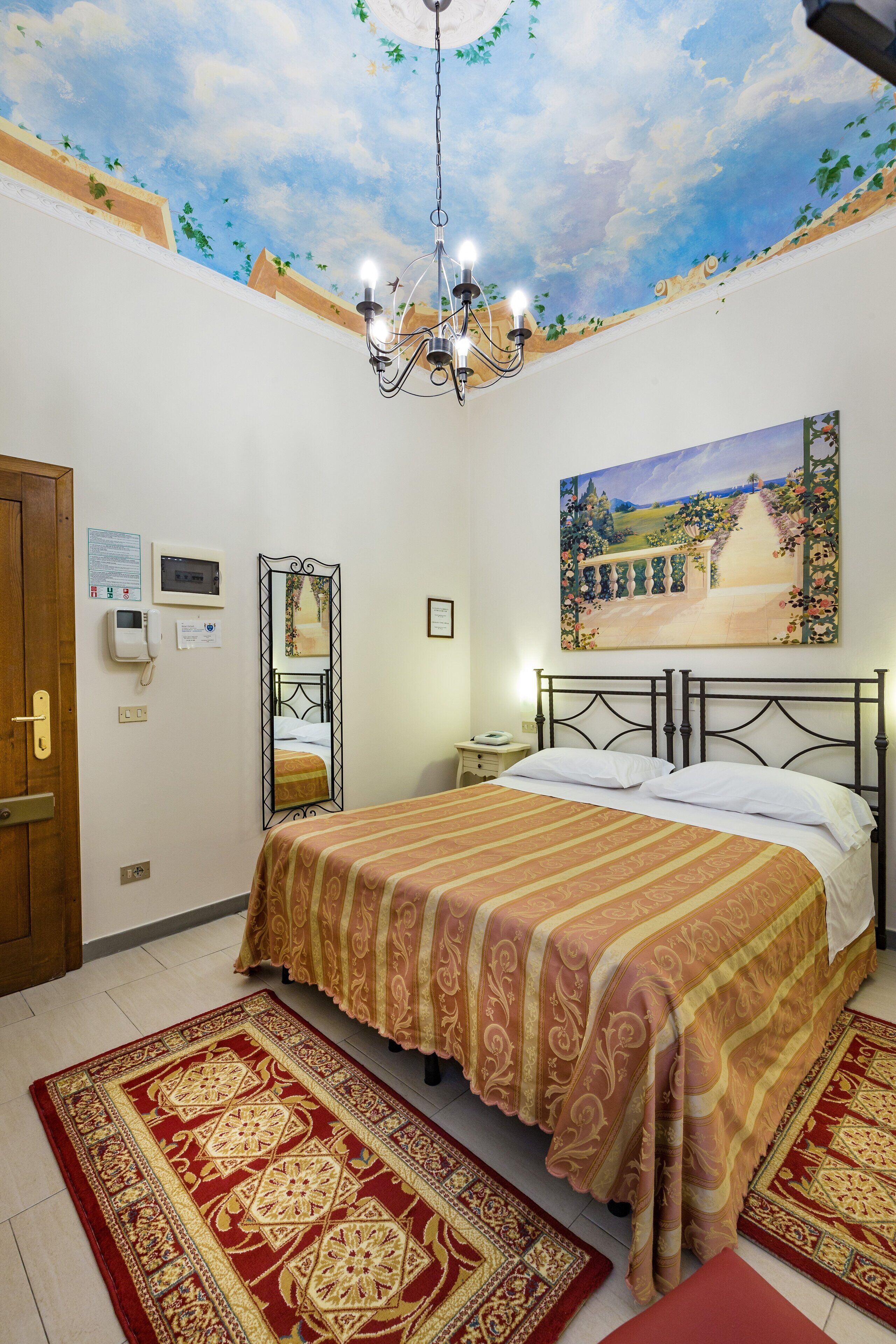 A bedroom with a bed, a chandelier, a painting, a mirror, and a carpet.