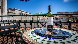 Una mesa con una botella de vino y dos copas sobre un mantel de mosaicos, ubicada en un balcón con vistas a edificios.