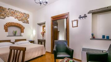 Una camera d'albergo con un letto, due poltrone, un tavolo con una ciotola di frutta, uno specchio, un muro in pietra e un lampadario.