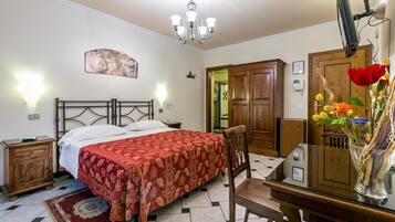 A bedroom with a bed, a nightstand, a desk with a chair, a wooden wardrobe, and a chandelier.