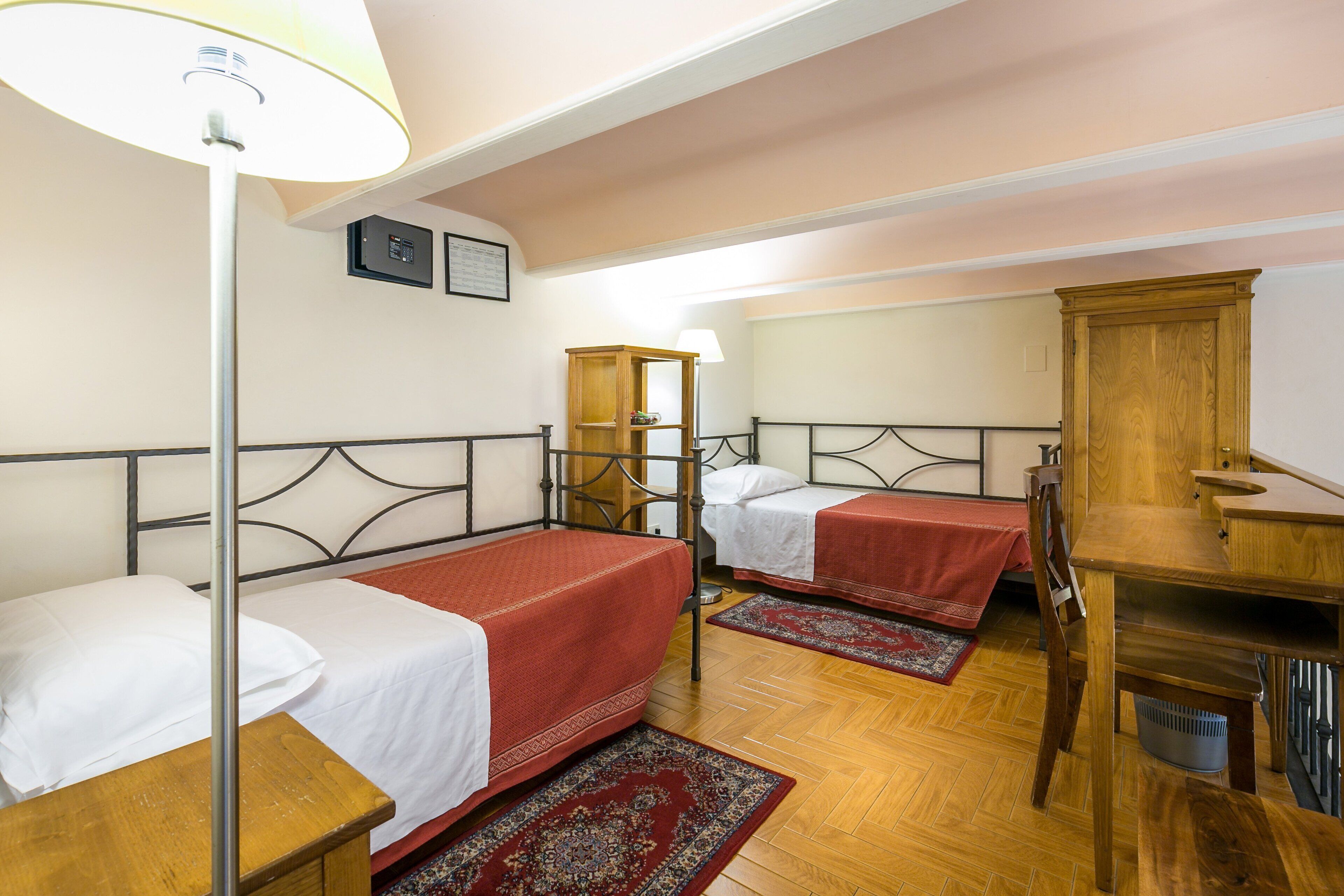 A room with two single beds, a wooden desk, a chair, a lamp, and a small cabinet.