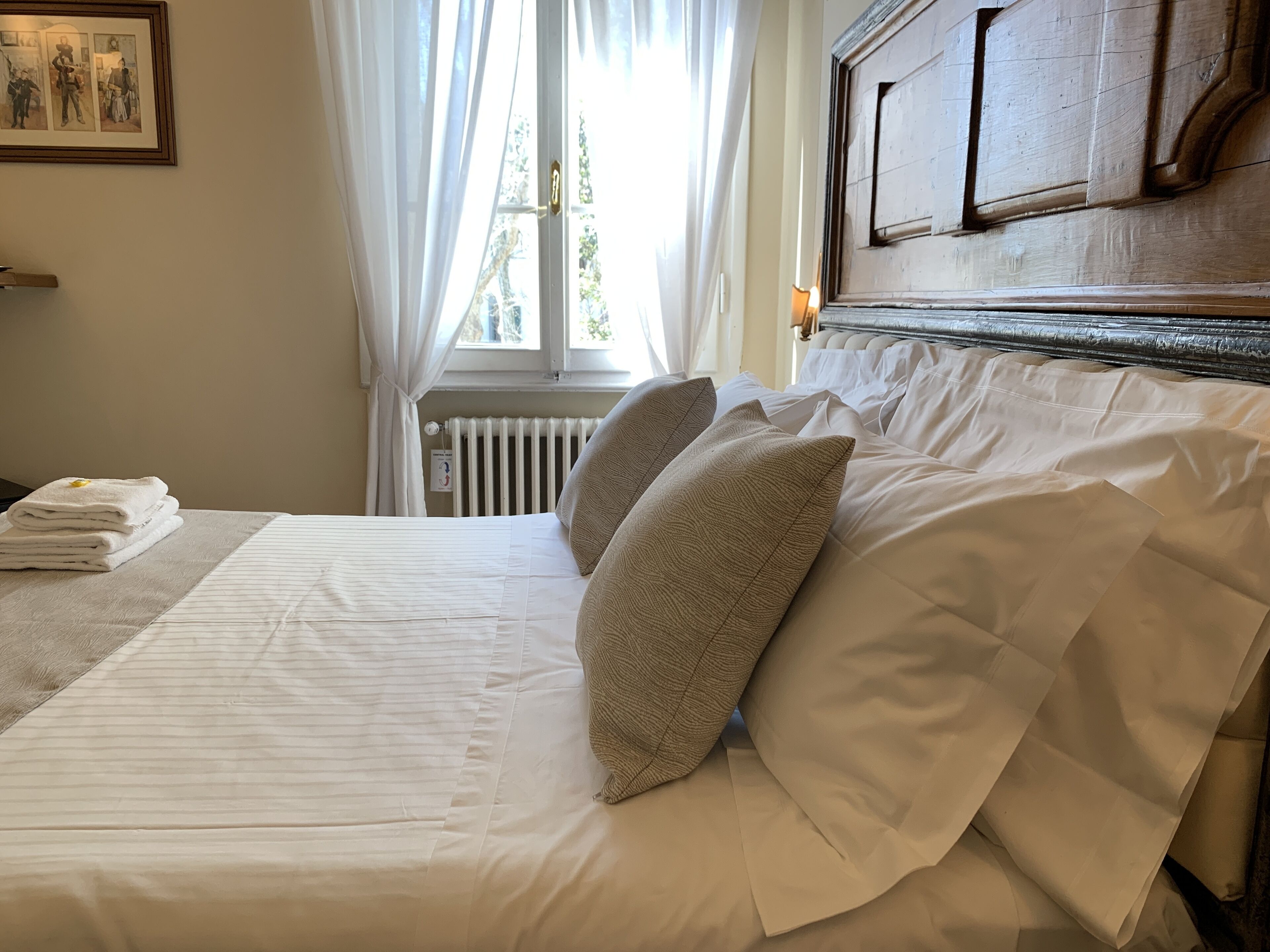 Una cama bien hecha con sábanas blancas y dos almohadas color beige, un radiador y un cuadro en la pared.