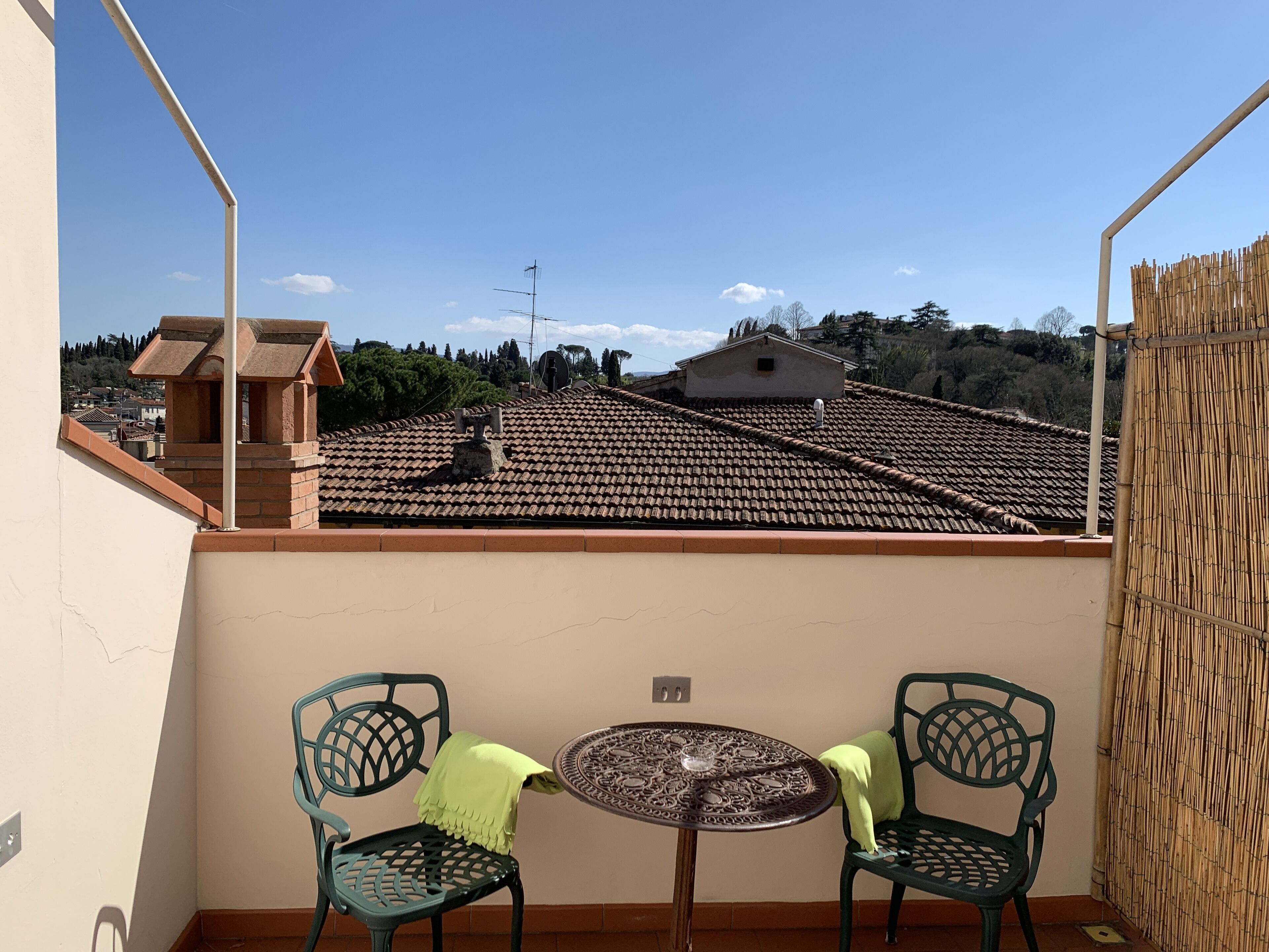 A balcony with two green chairs and a small table, overlooking a tiled roof and distant buildings.