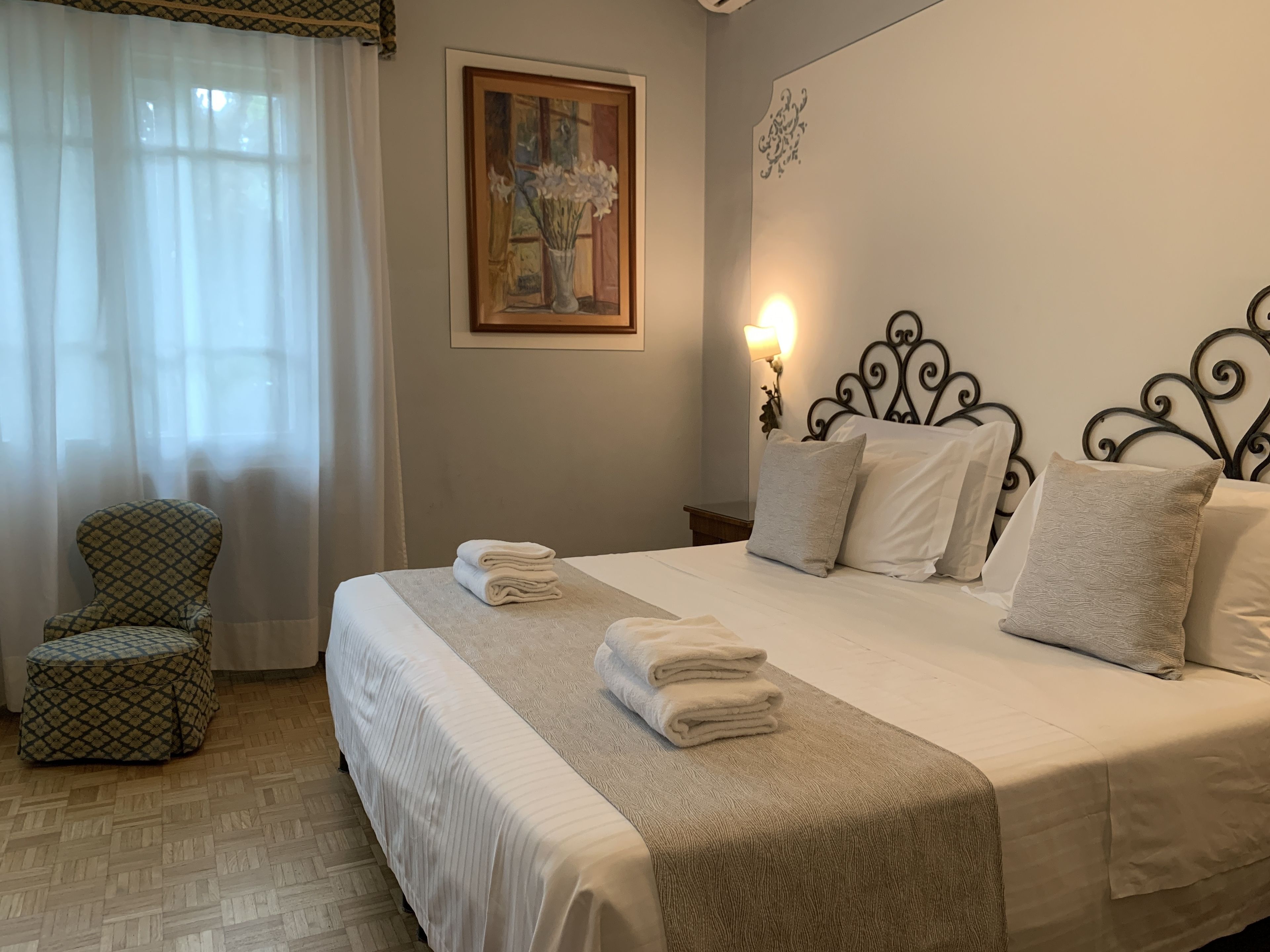 A neatly made bed with white linens and a beige throw blanket, a patterned armchair, and a painting on the wall.