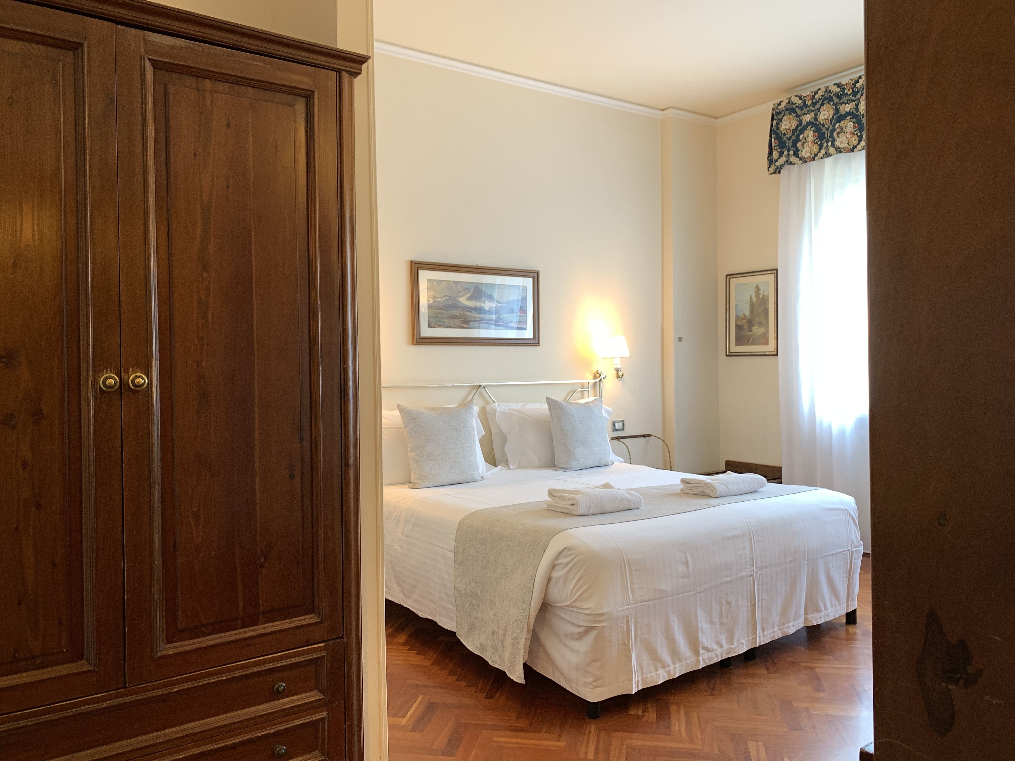 Habitación de hotel con armario de madera, cama con ropa blanca, mesita de noche y lámpara de pared.