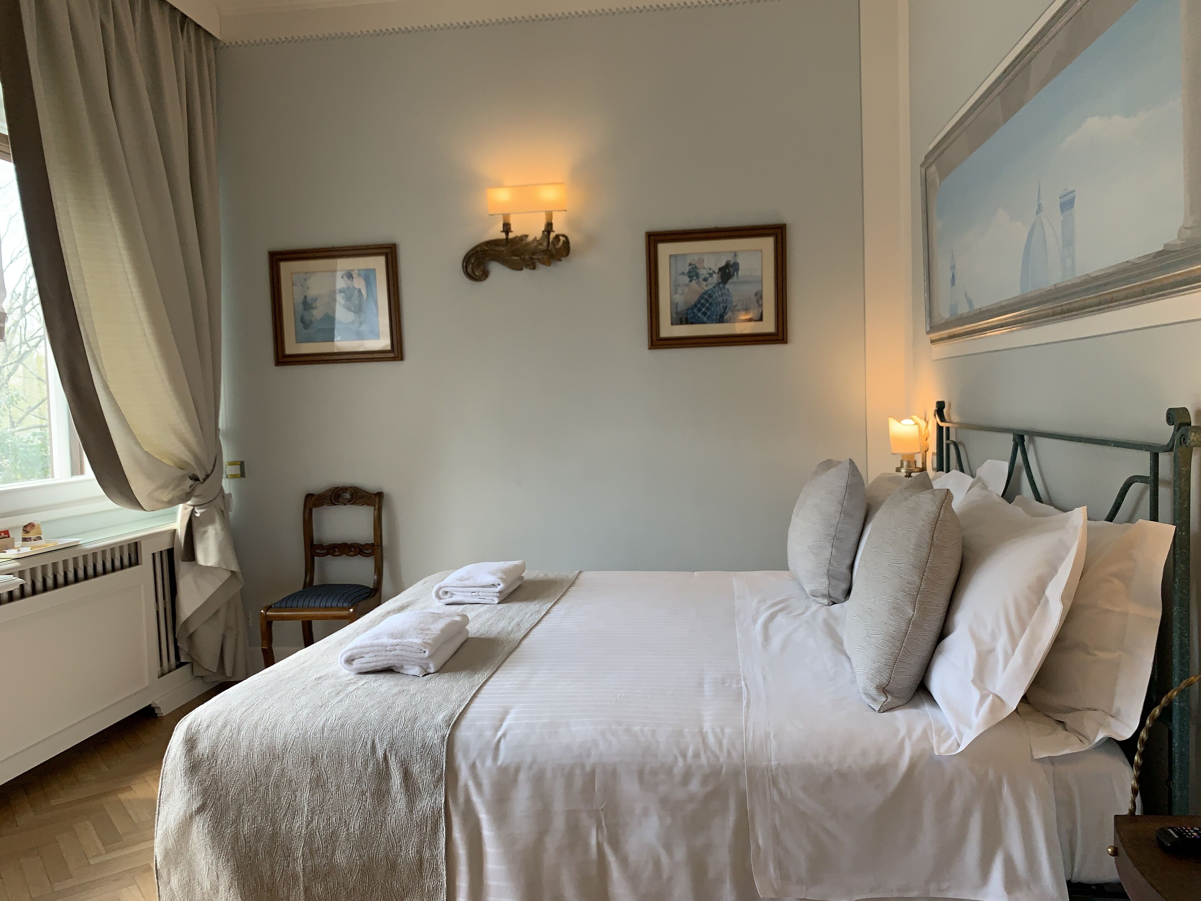 A neatly made bed with pillows and a folded towel, a chair, a radiator, and two wall-mounted light fixtures.