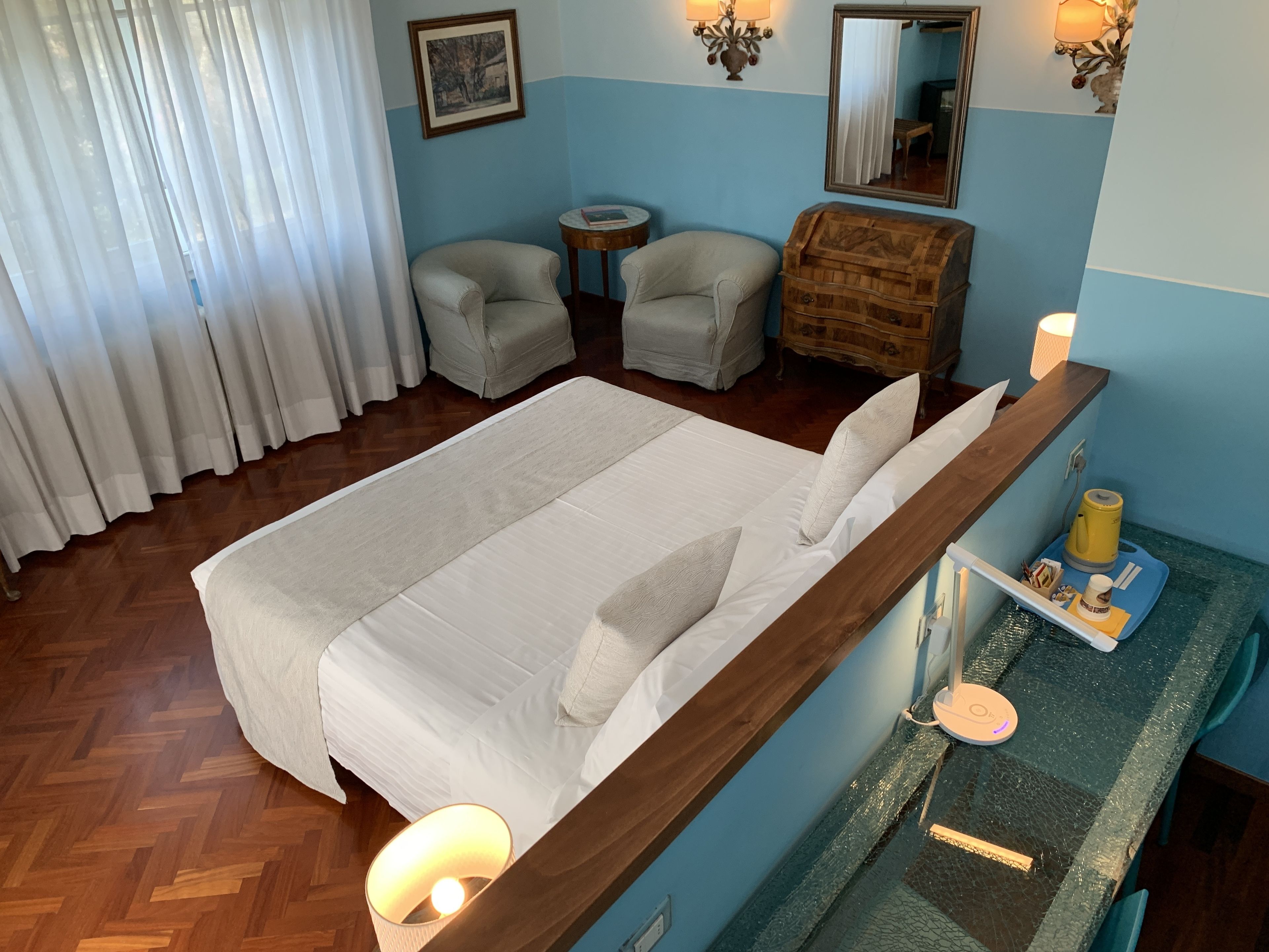A hotel room with a large bed, two armchairs, a small round table, a dresser, and a vanity with a mirror.
