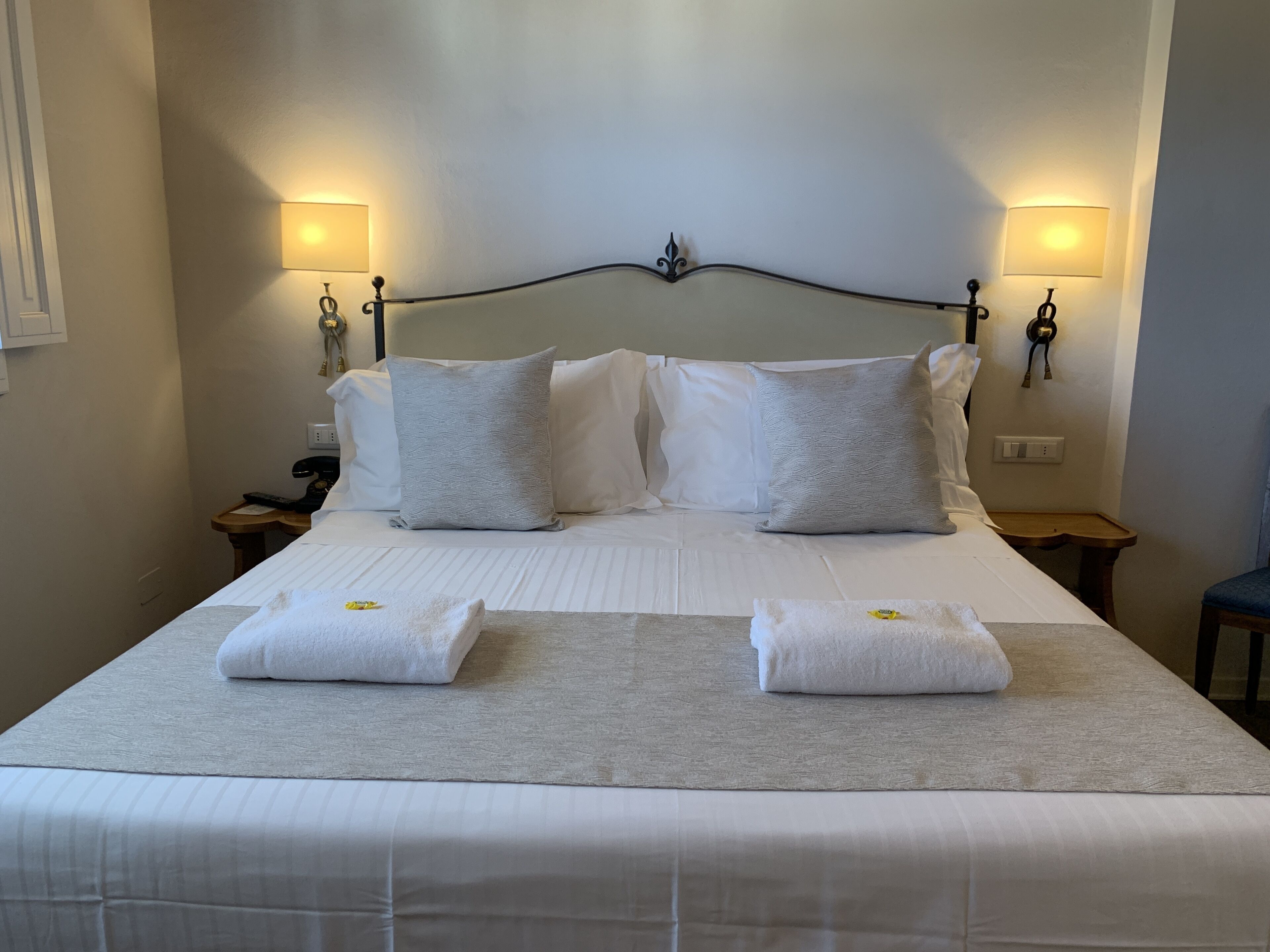 A neatly made bed with white linens and two pillows, two bedside tables with lamps, and a wall-mounted light fixture.