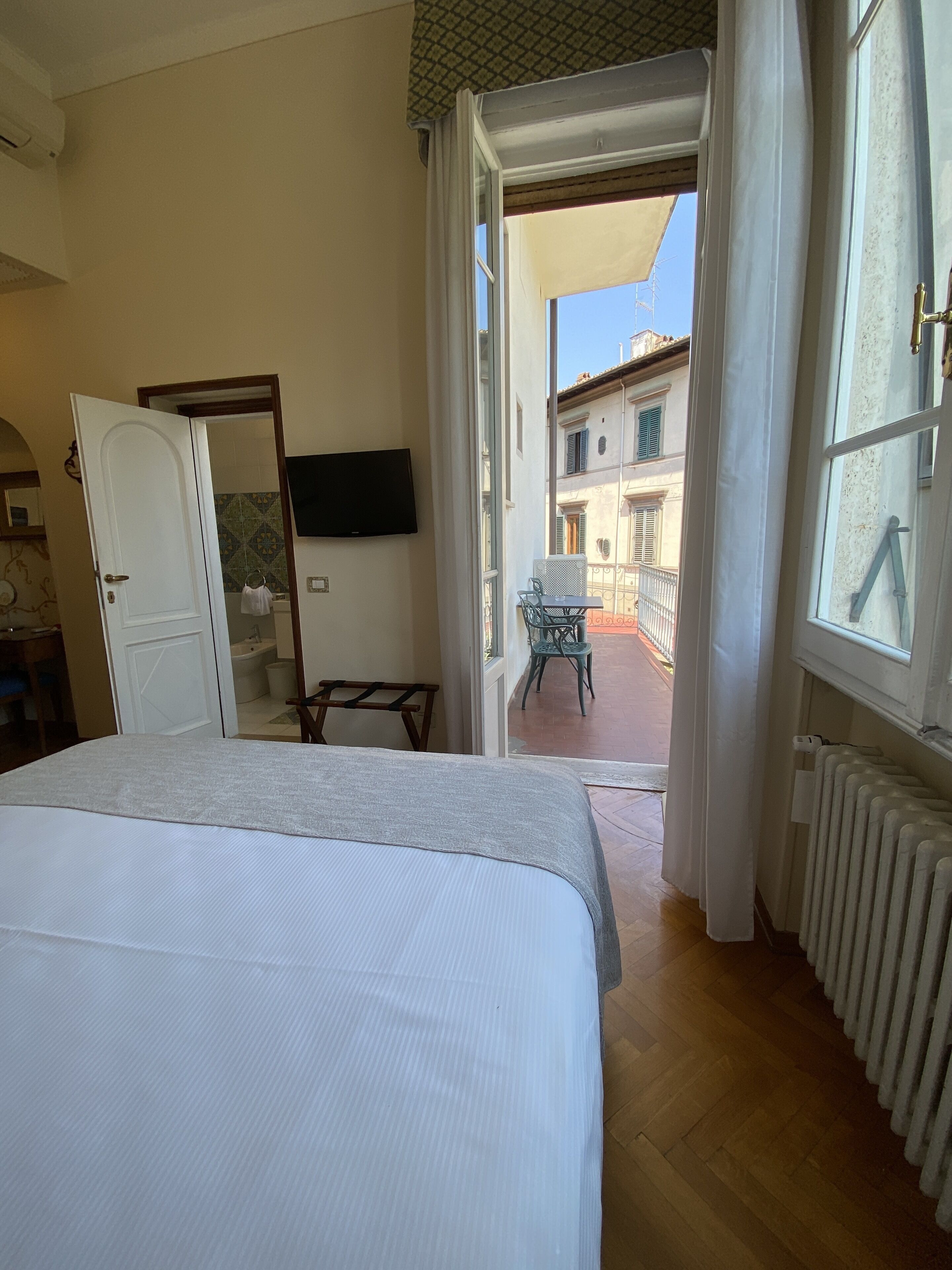 Une chambre à coucher avec un lit, une télévision et une vue sur une cour intérieure, où se trouvent une chaise et une table.