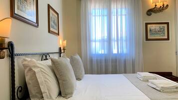 Una cama bien hecha con sábanas y almohadas blancas, una mesita de noche con lámpara, y una ventana con cortinas traslúcidas que dejan entrar luz natural.