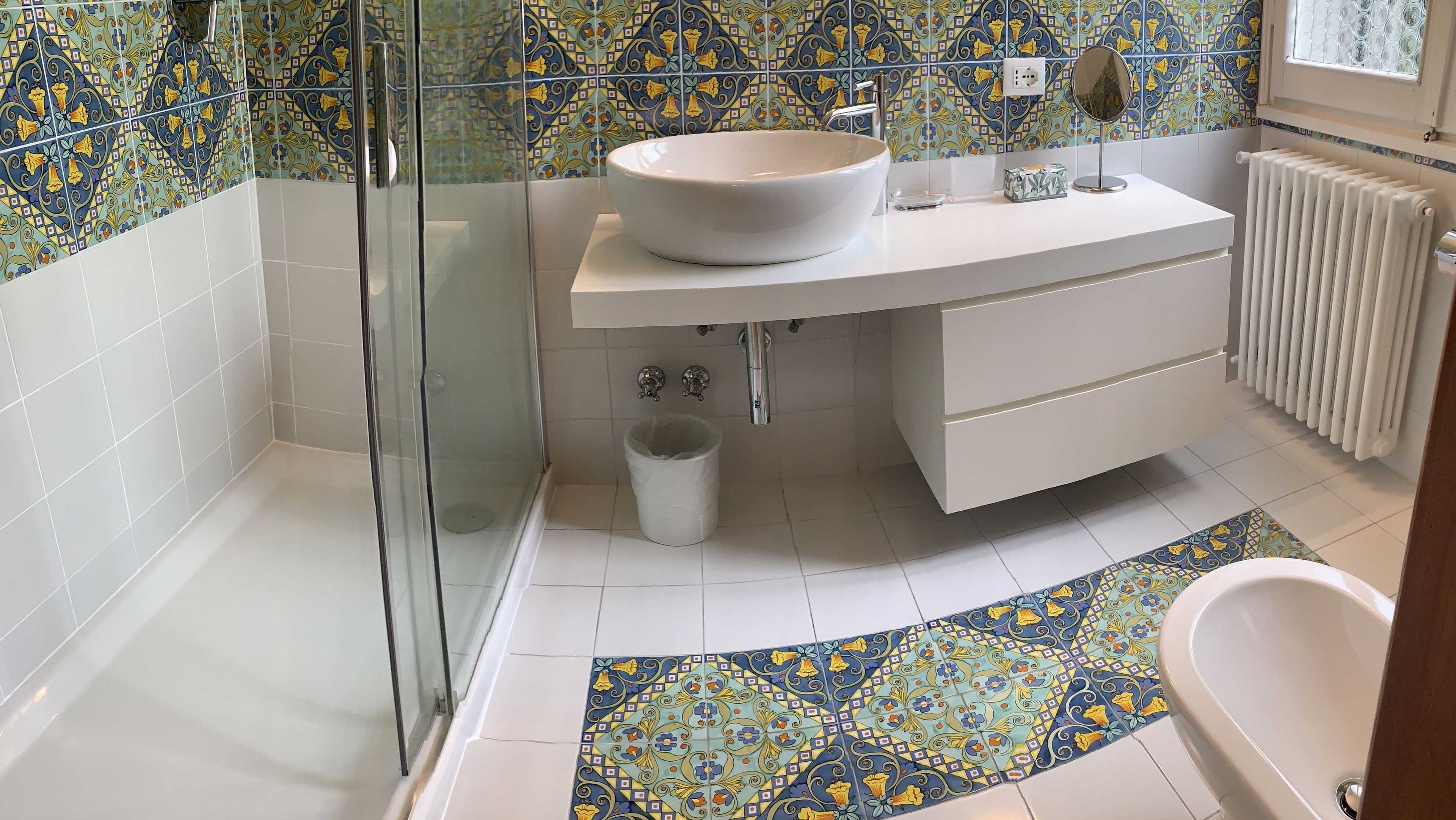 Un baño con ducha envuelta en vidrio, un lavabo blanco con pileta a la vista, un mueble de tocador blanco y un piso con diseño.
