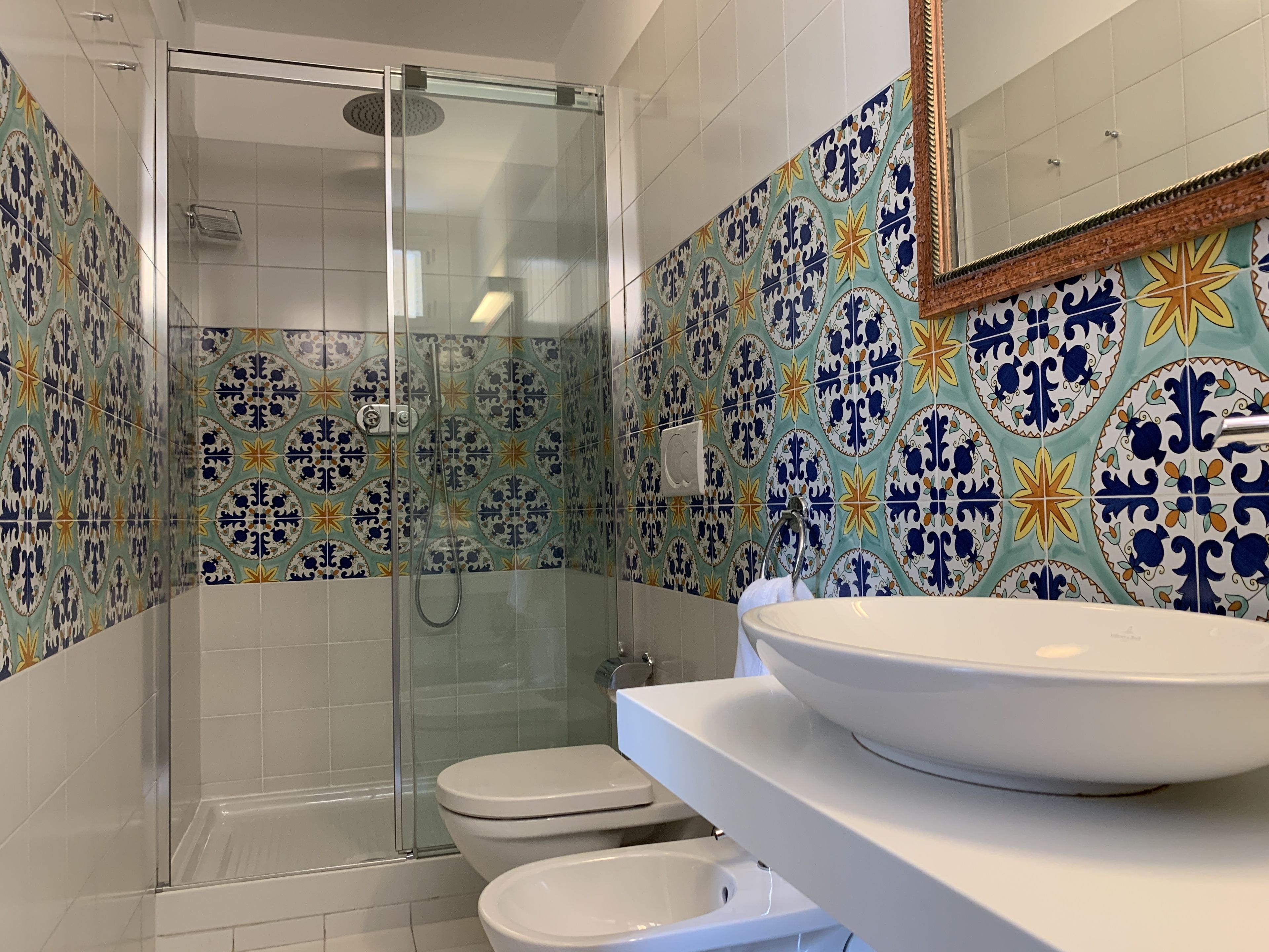 Un bagno con doccia vetrata, un lavandino bianco con una ampia bacinella e un water. Le pareti sono decorate con piastrelle colorate e a motivi.