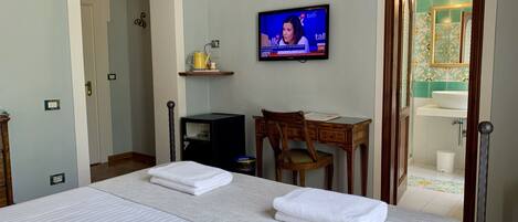 Ein Hotelzimmer mit Bett, Schreibtisch, Stuhl, Fernseher und einem durchgegangenen Tür sichtbar liegenden Badezimmer.