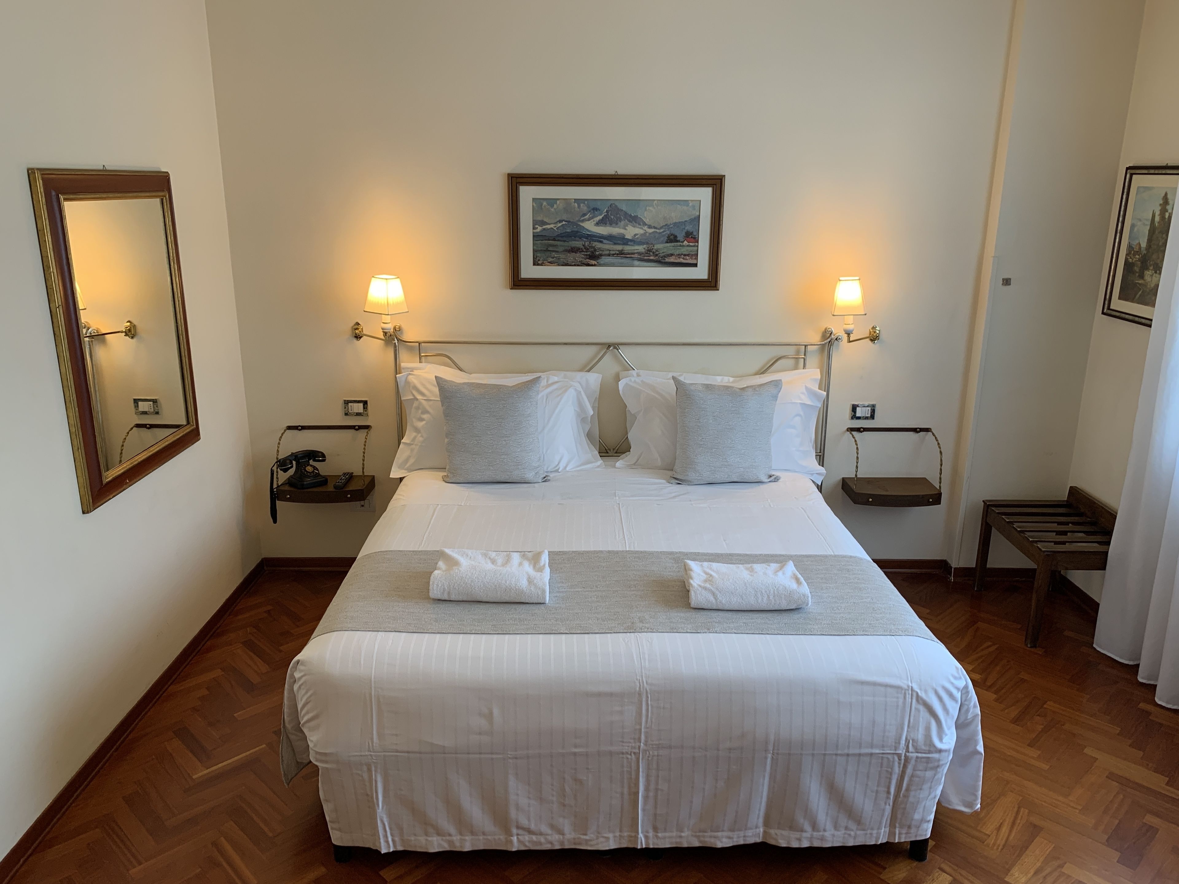 A neatly made bed with white linens and two pillows, a bedside lamp, a framed picture on the wall, and a wooden bench.