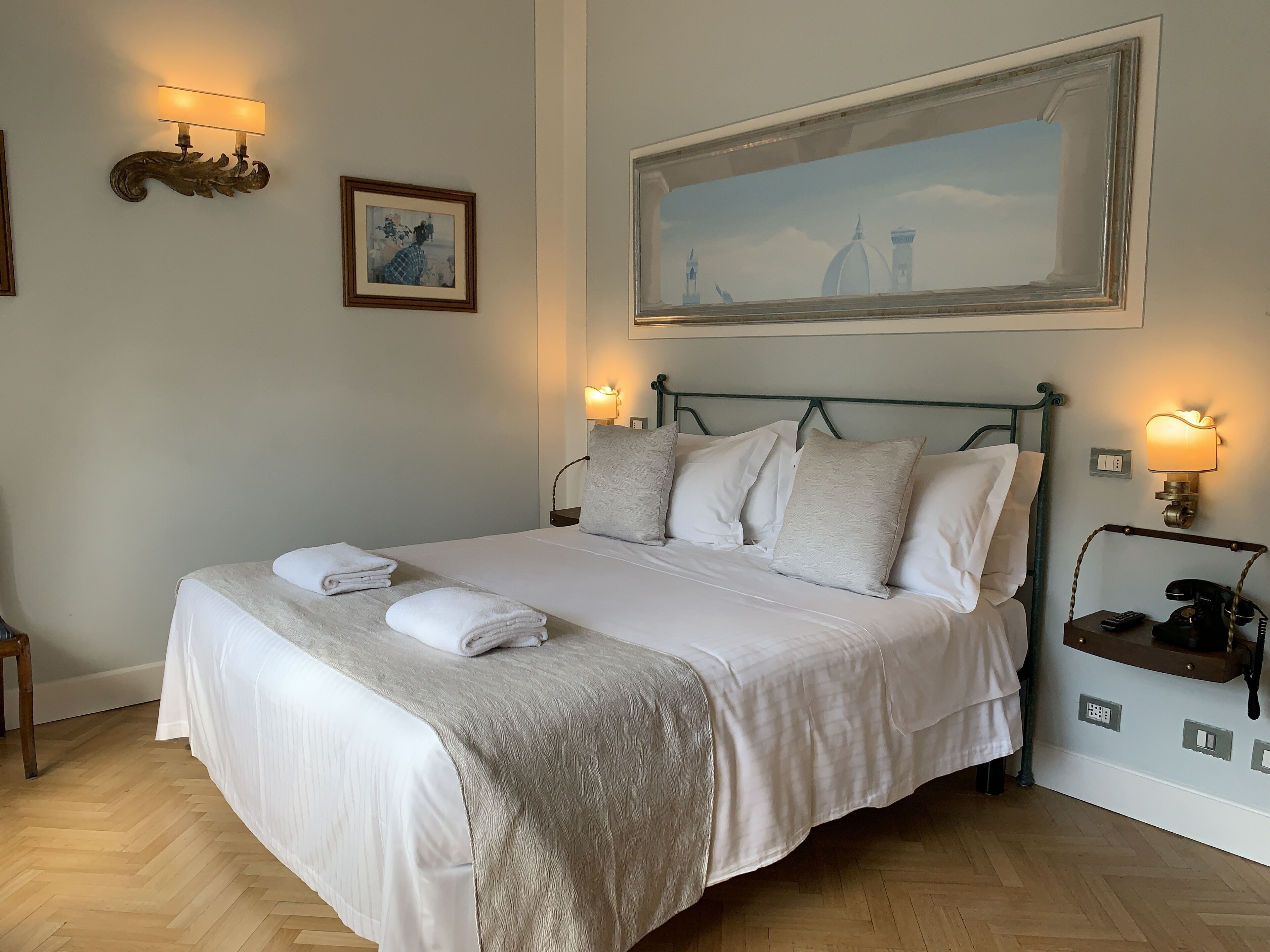 A neatly made bed with white linens and a beige throw blanket, two wall-mounted lamps, a framed painting, and a large landscape painting above the bed.