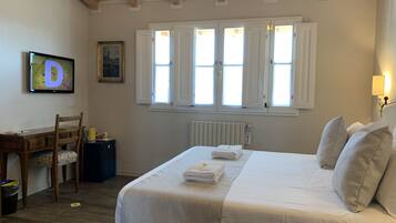 A bedroom with a wooden ceiling, a bed with white linens, a desk with a chair, and a window with white shutters.