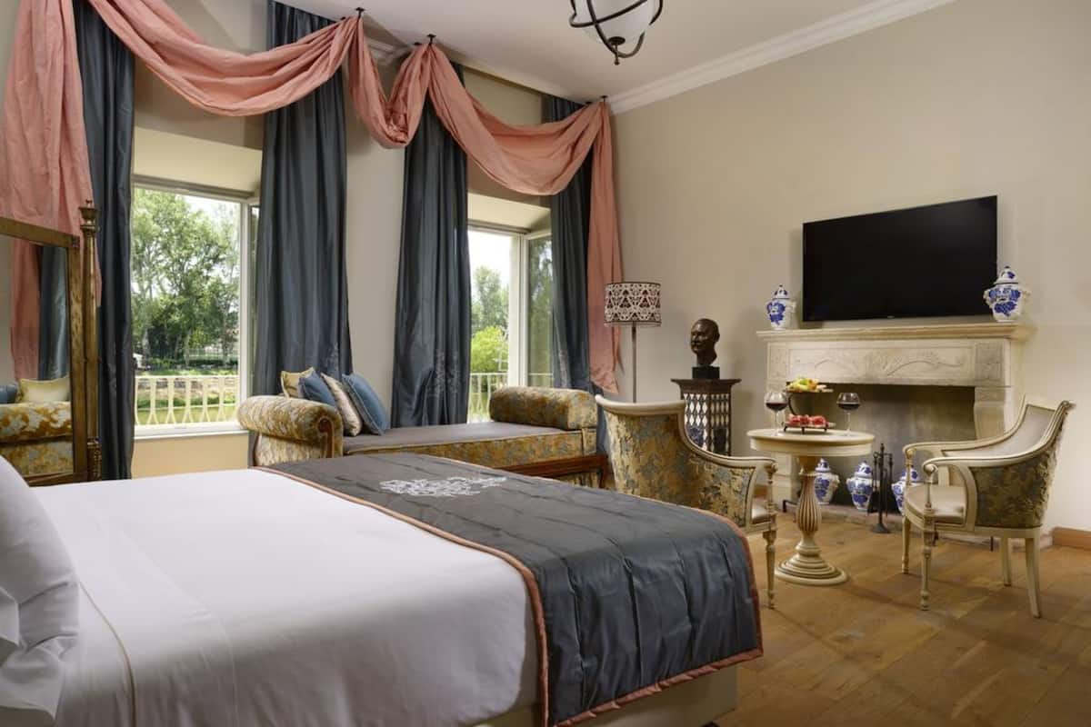 A hotel room with a large bed, a sofa, a fireplace, a television, and a view of the outdoors.