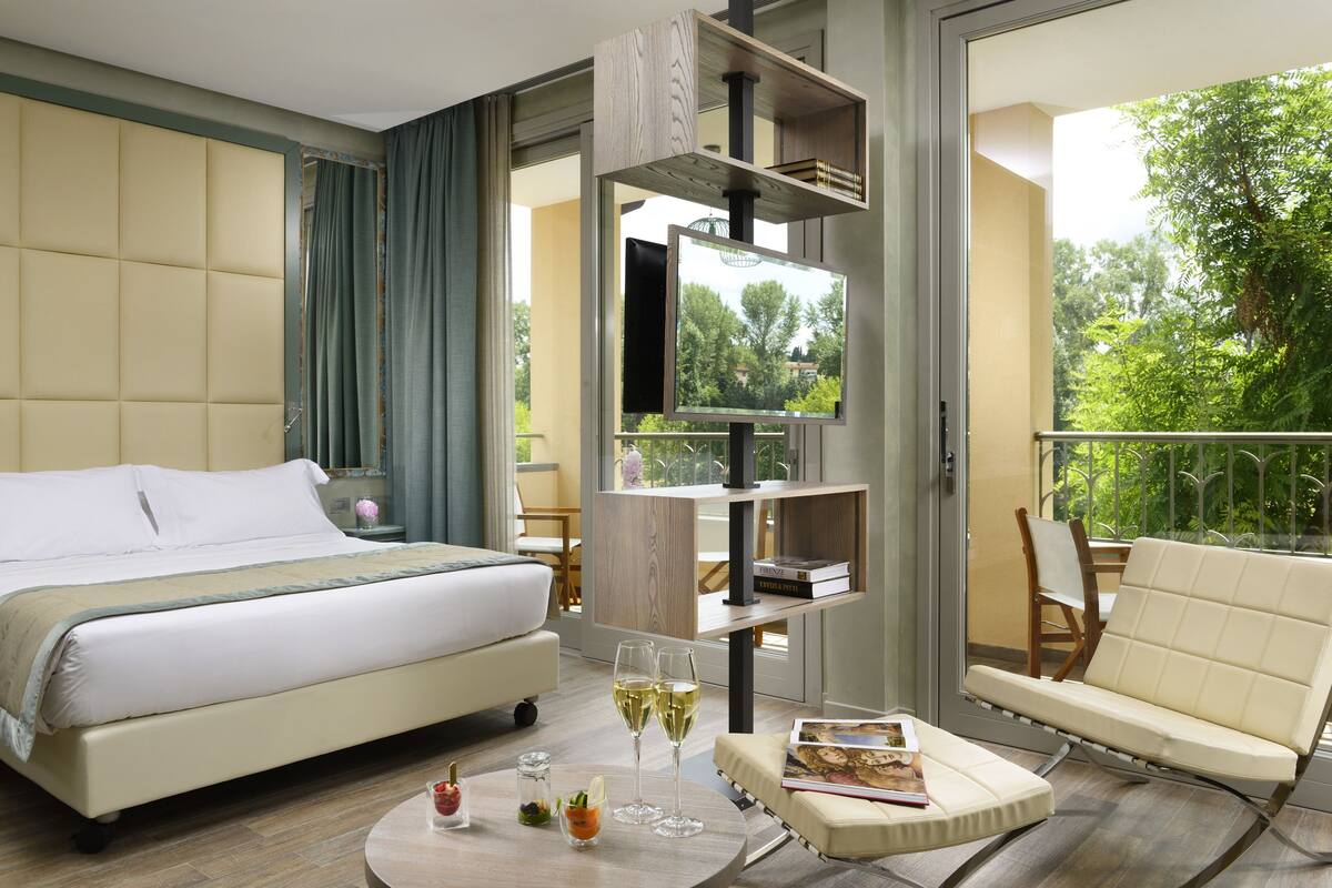 A hotel room with a large bed, a sofa, a round coffee table with glasses and snacks, a bookshelf, and a balcony with a view of trees.