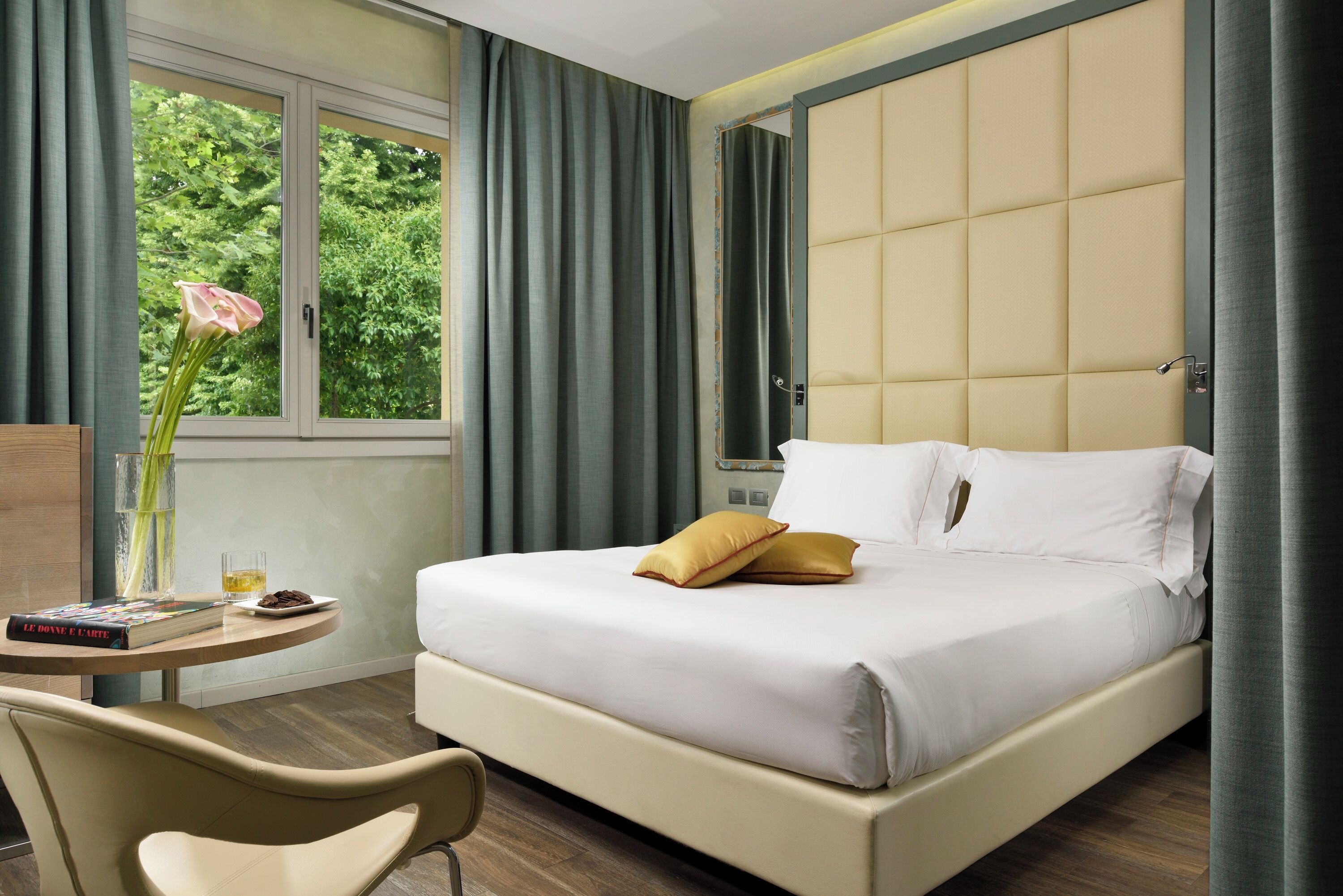Une chambre d’hôtel avec un lit, une table de chevet sur laquelle se trouvent un livre et un verre, une chaise et une fenêtre avec des rideaux.