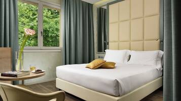 Habitación de hotel con cama, mesita de noche con un libro y un vaso, una silla y una ventana con cortinas.
