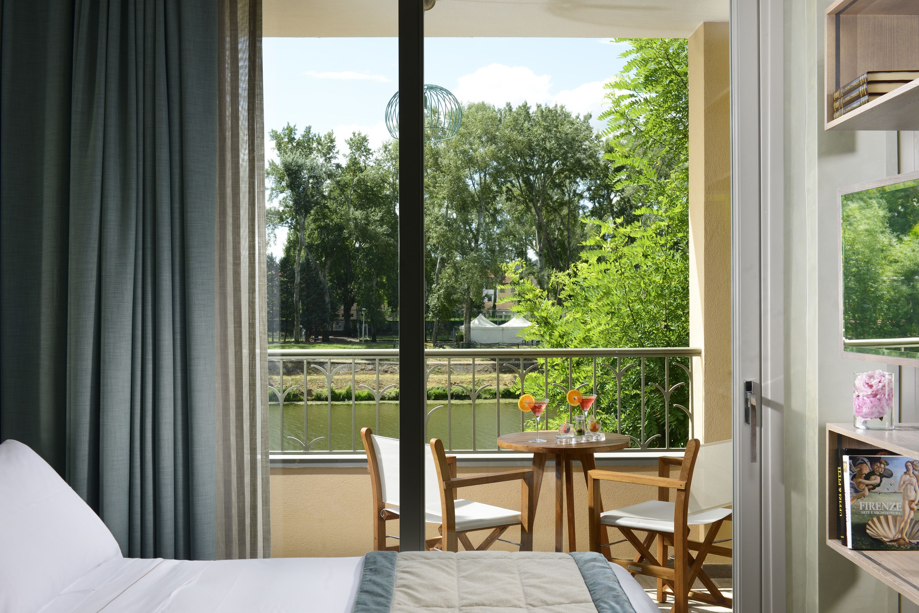 A hotel room with a balcony, a bed, a table with chairs, and a view of a river and greenery.