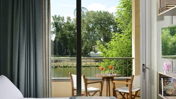 A hotel room with a balcony, a bed, a table with chairs, and a view of a river and greenery.
