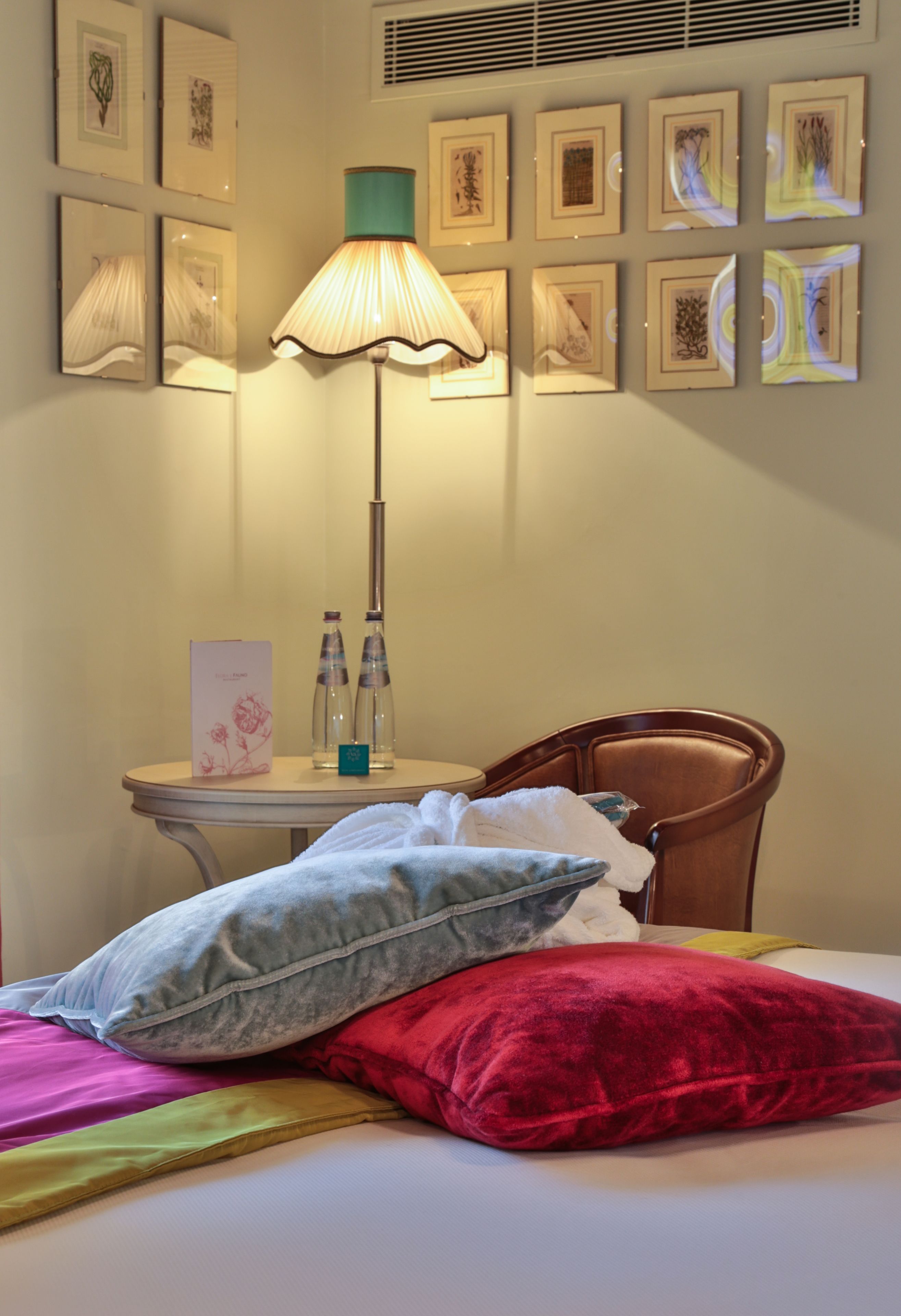 Een hotelkamer met een bed, kussens, een tafel met waterflesjes, een lamp en ingelijste foto's aan de muur.